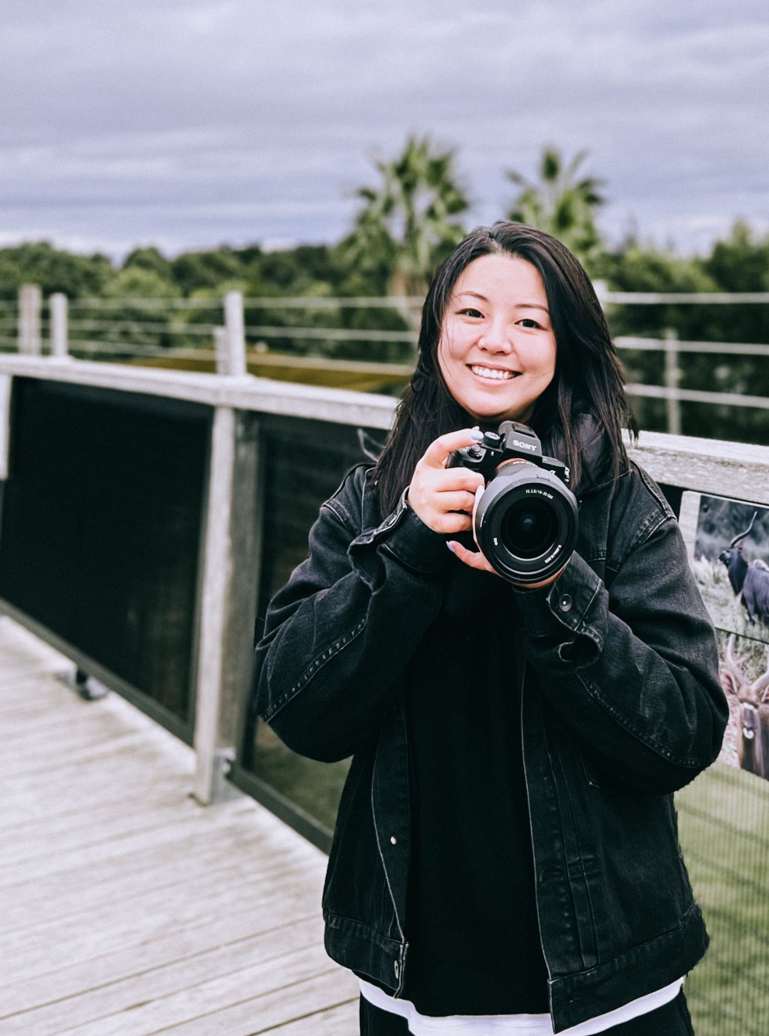 A photographer in a black jacket holds a professional DSLR camera while standing on an outdoor deck with palm trees visible.