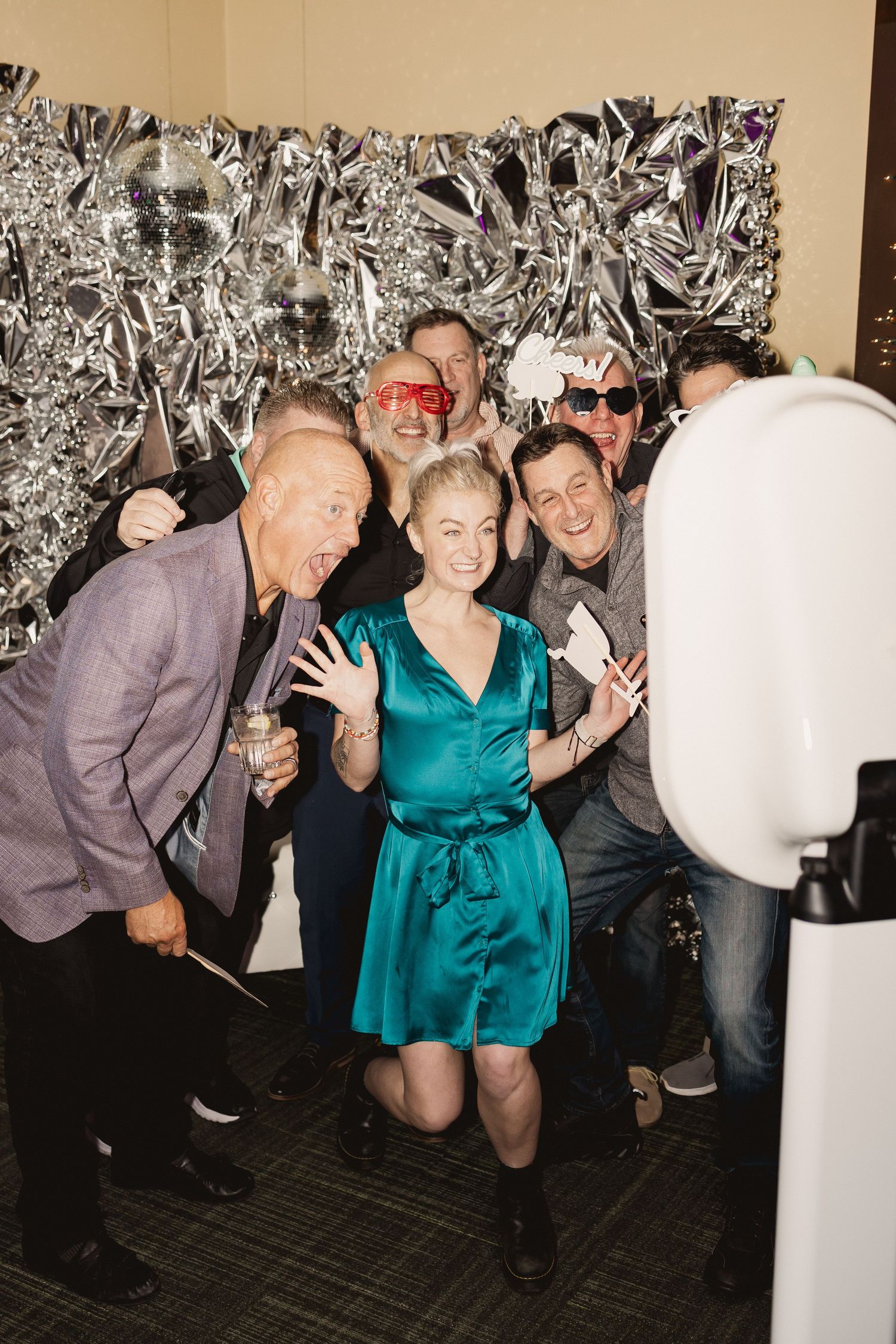 A group of party guests gather around a photo booth against a silver backdrop at a celebration.