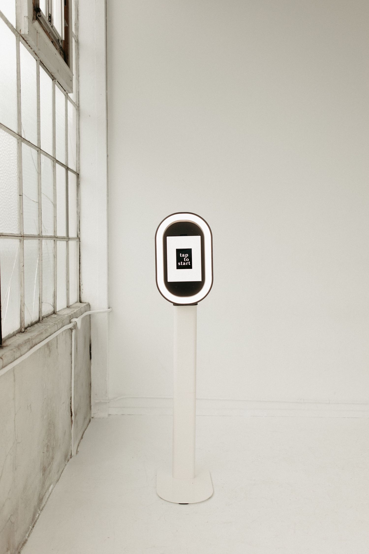 A minimalist white photo booth kiosk with a digital display screen stands against a window in a bright room.