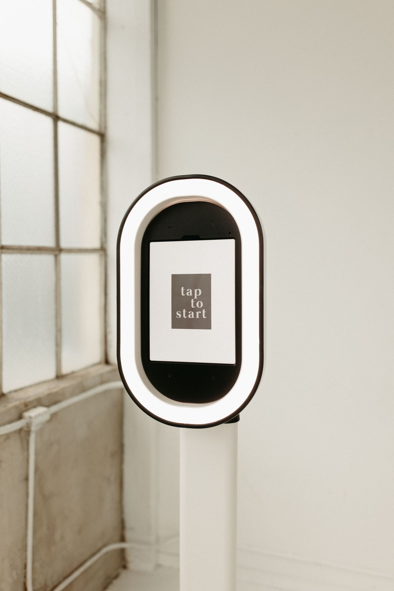 Close-up of a sleek photo booth display screen mounted on a white stand near a window.
