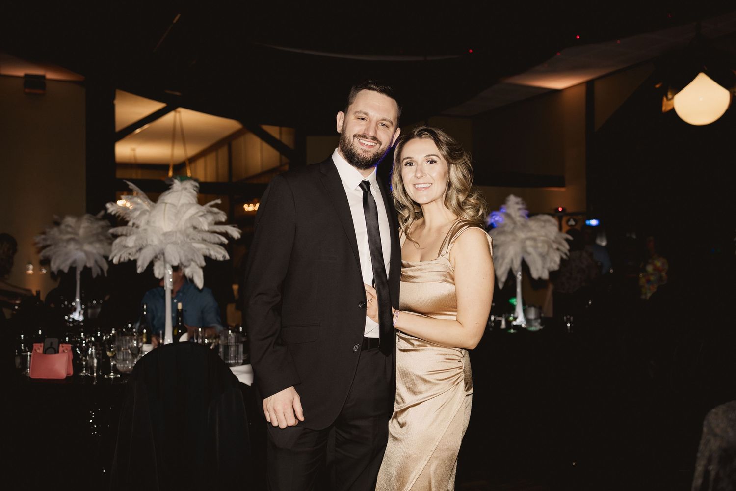 Couple in formal attire poses at event with feather arrangements in background.