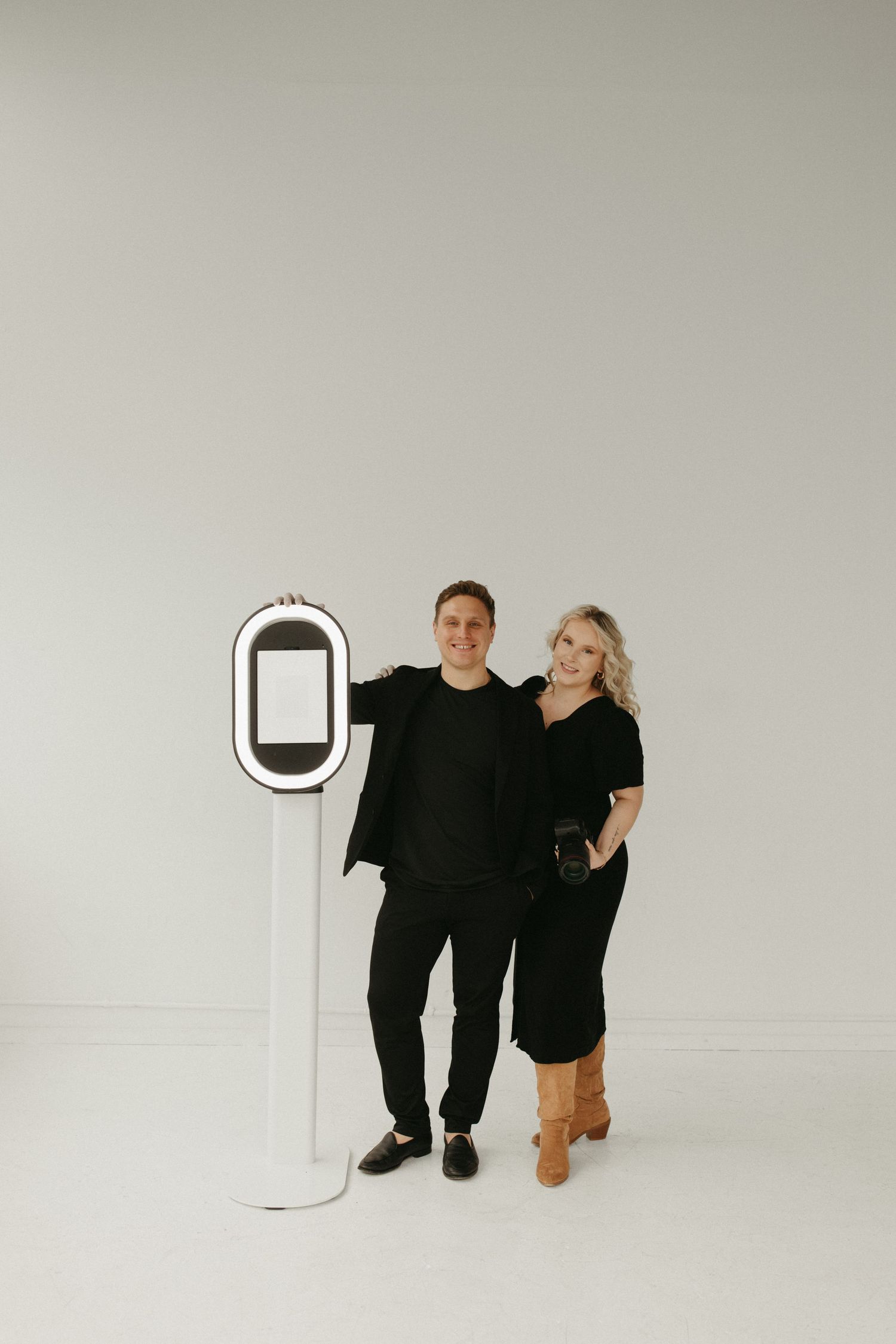 Minimalist studio portrait with two people standing beside white digital kiosk against neutral background.