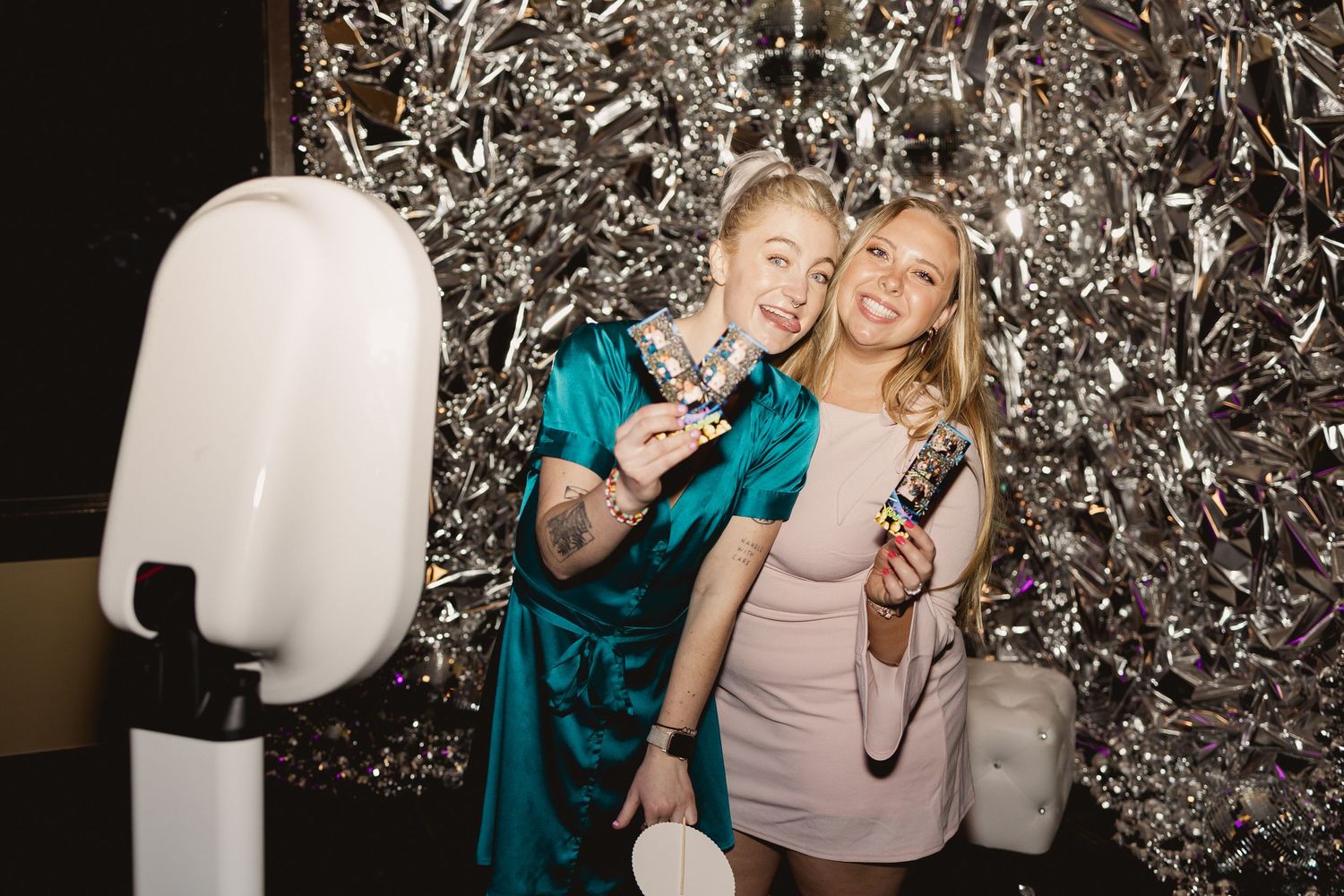 Two women pose with drinks in front of a silver photo backdrop.