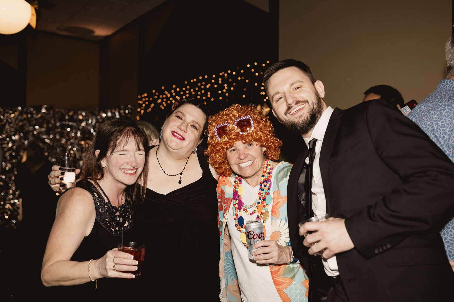 Friends in festive attire smile together at party with sparkly backdrop.