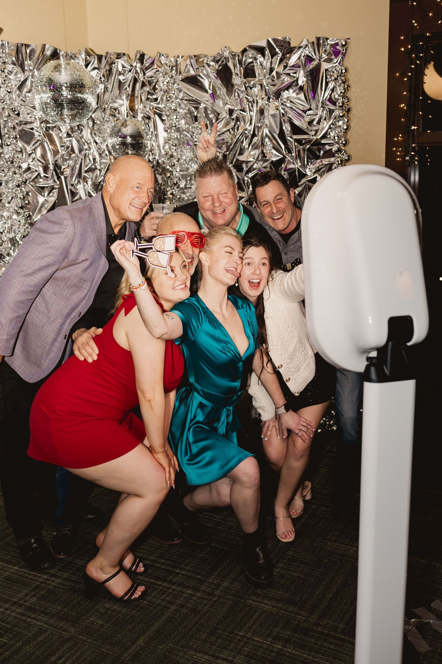 A group poses playfully in front of a silver backdrop at a party photo booth.