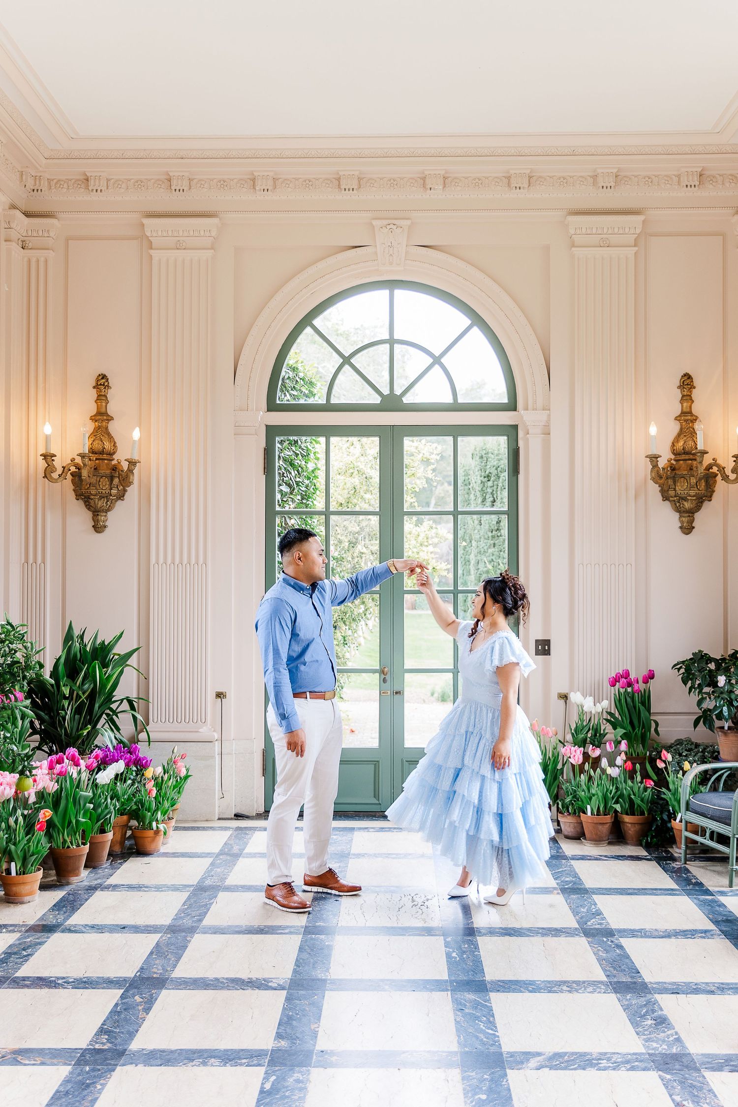 Couple dancing in elegant sunlit room with arched windows and pink tulips.