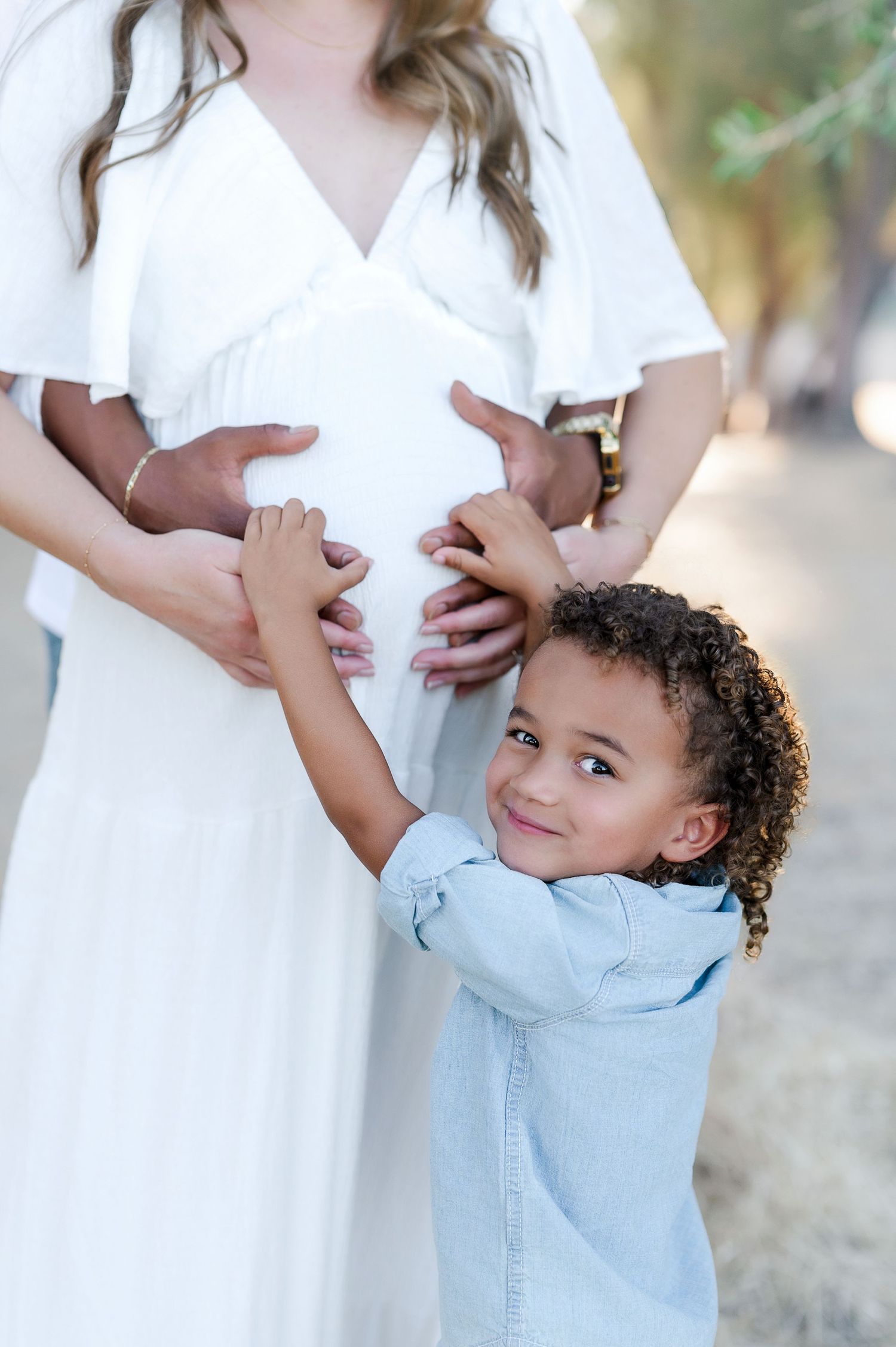 Young child hugs pregnant woman wearing white dress in outdoor setting.