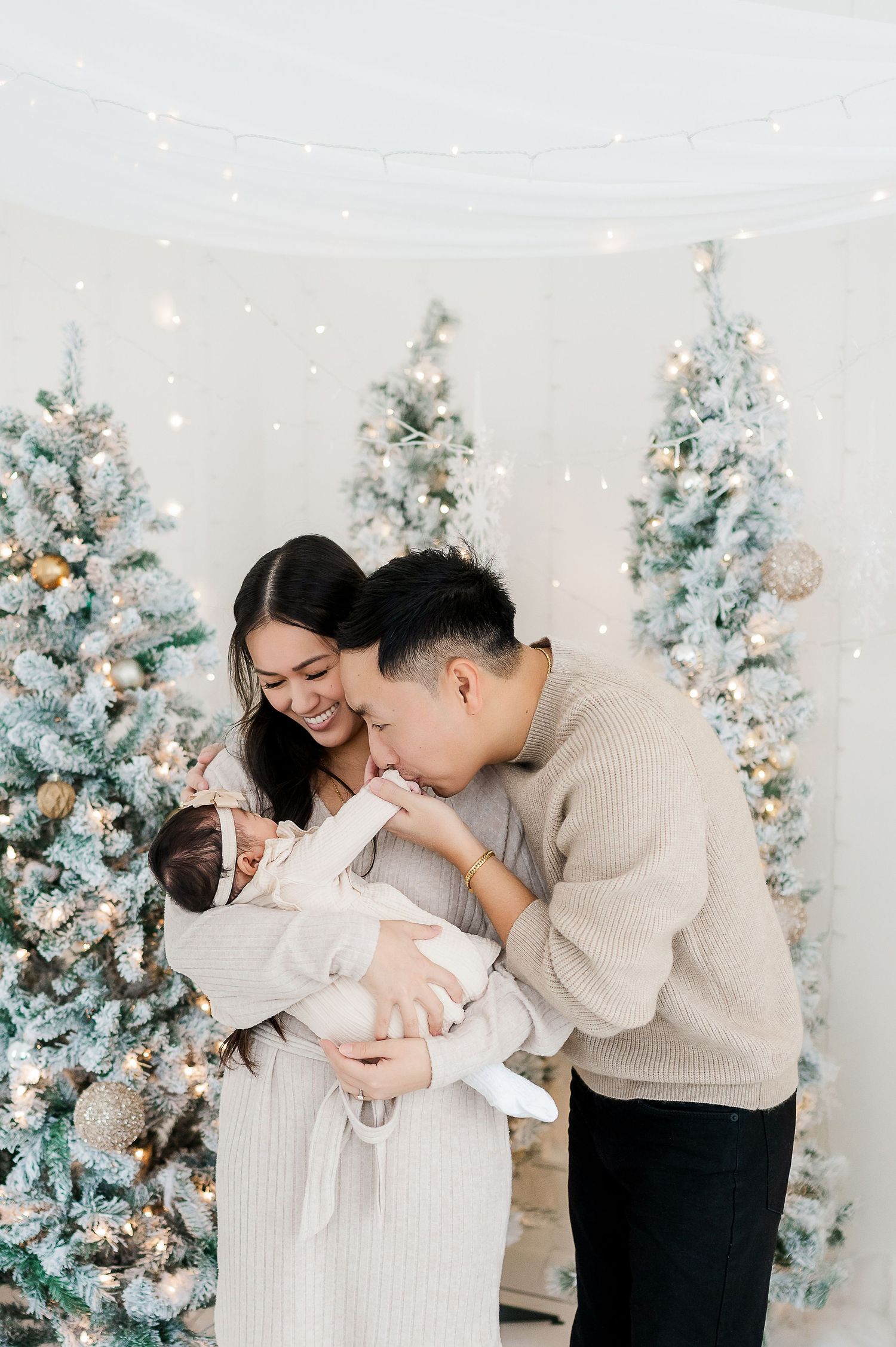 Family of three shares an intimate moment in front of decorated white Christmas trees.