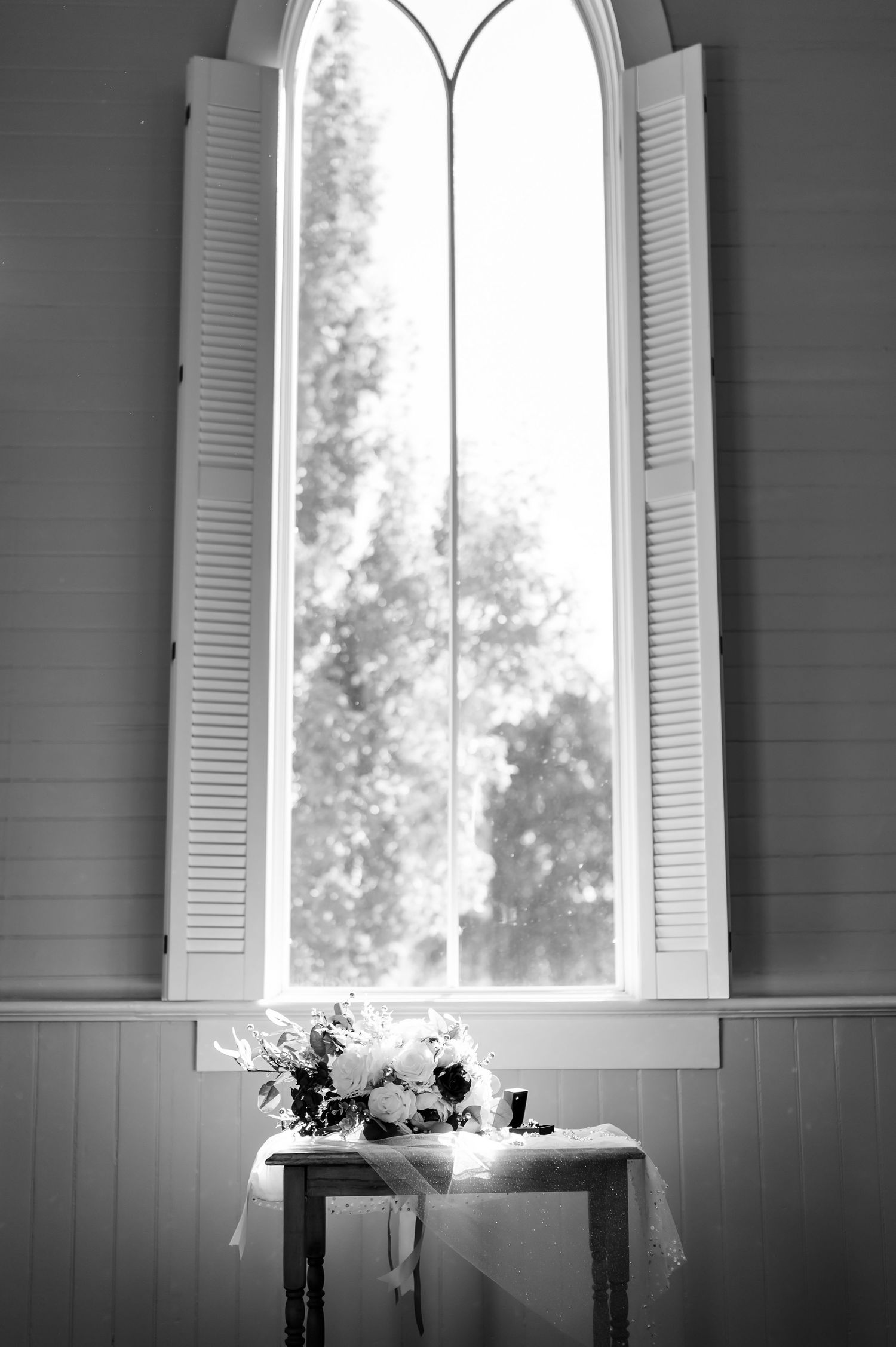 Black and white photo of an arched window with white shutters and small floral arrangement on table.