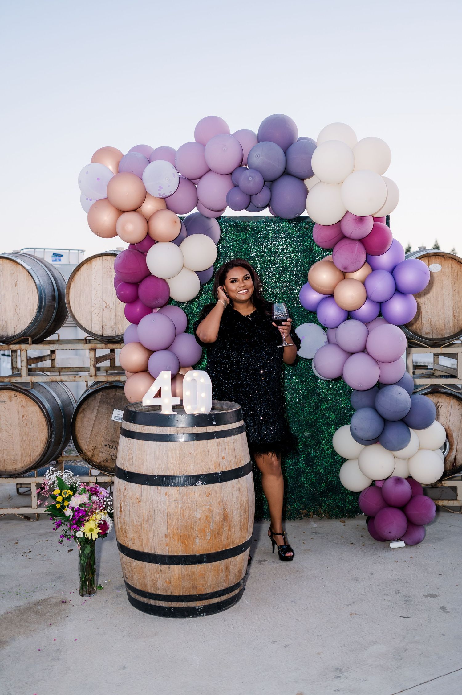 Birthday celebration setup with purple balloon arch and wine barrels in winery setting.