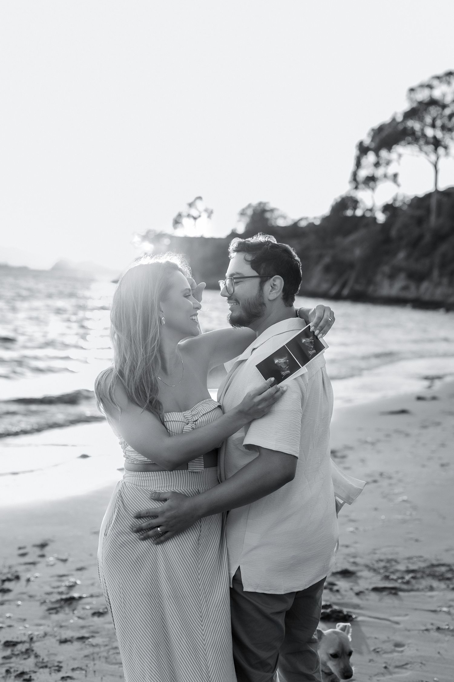 Black and white beach photo of couple embracing while holding ultrasound image.