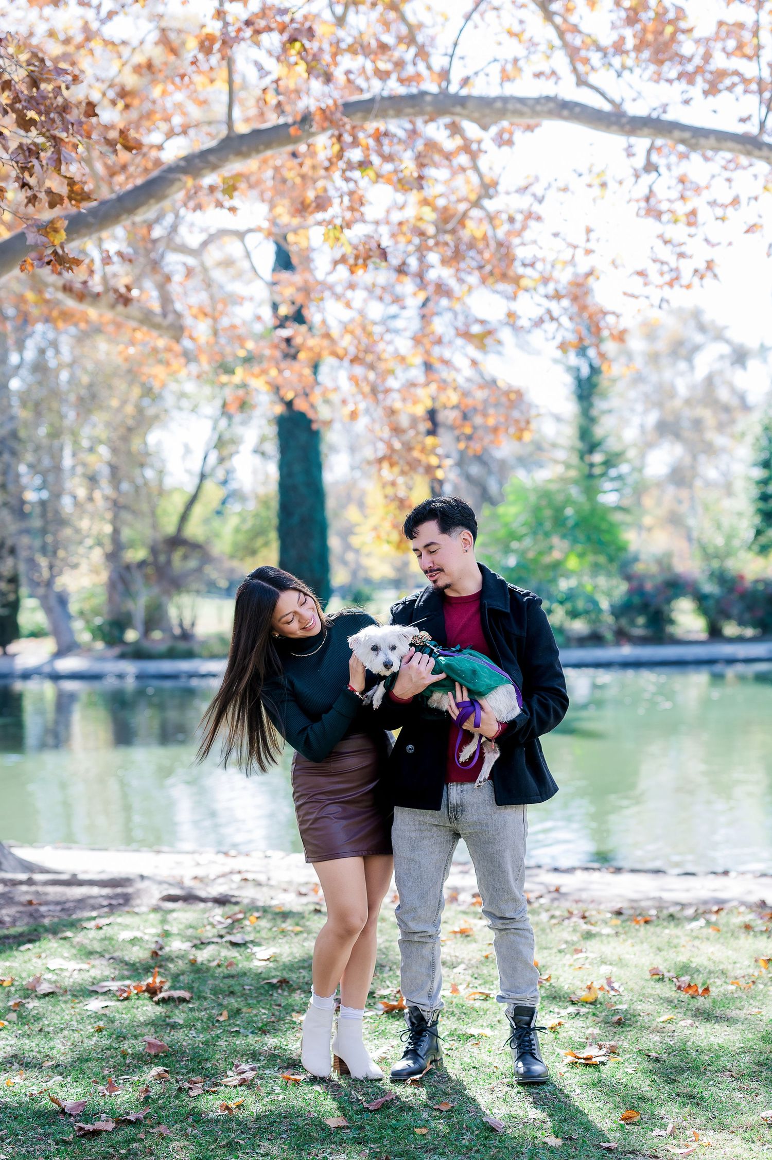 Couple holding small white dog while enjoying autumn scenery by a lake in a park.