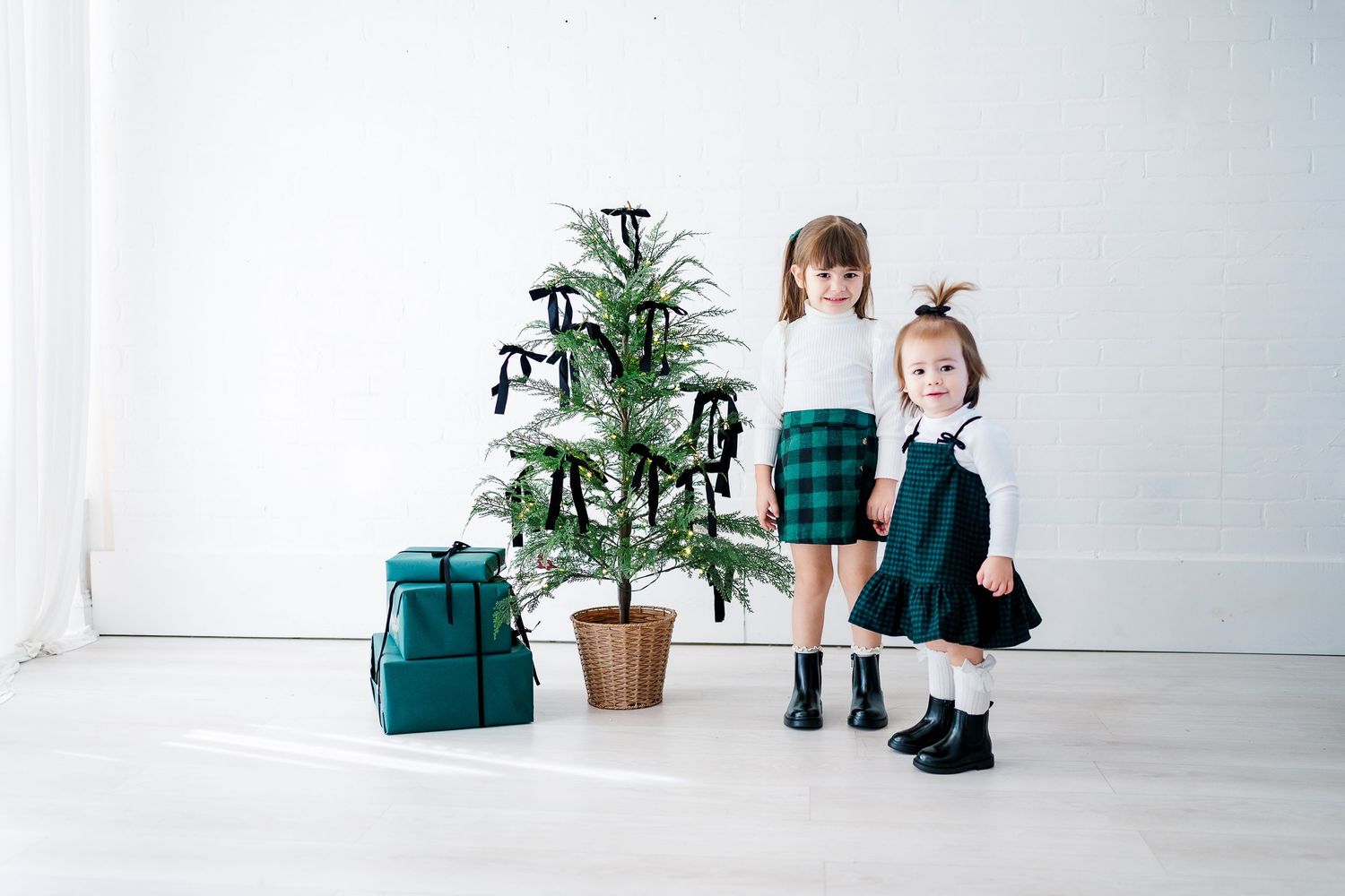 Two children in matching green outfits stand beside decorated Christmas tree with wrapped presents.