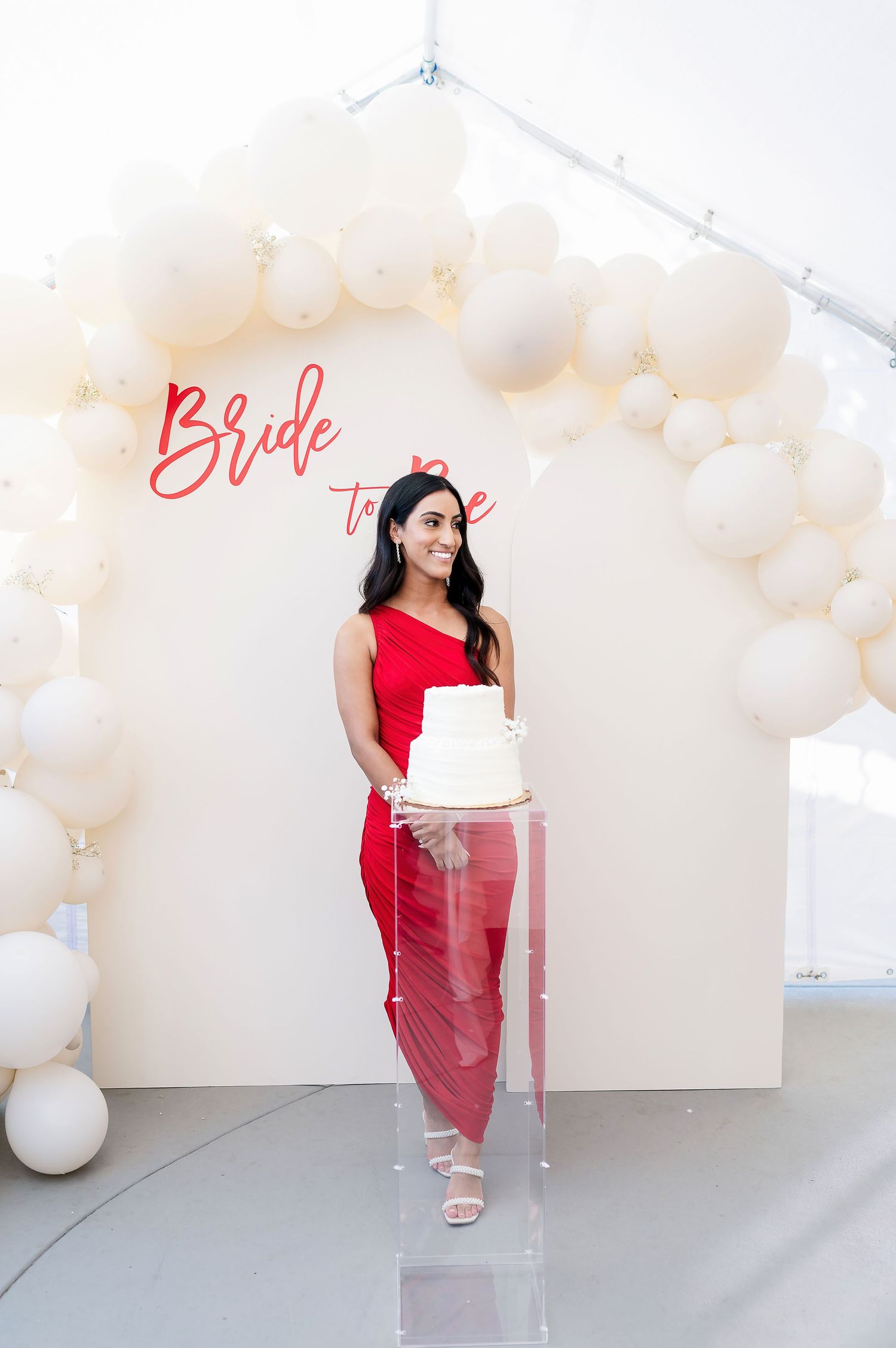 A bridal event setup with white balloon arch and acrylic display stand featuring a wedding cake.