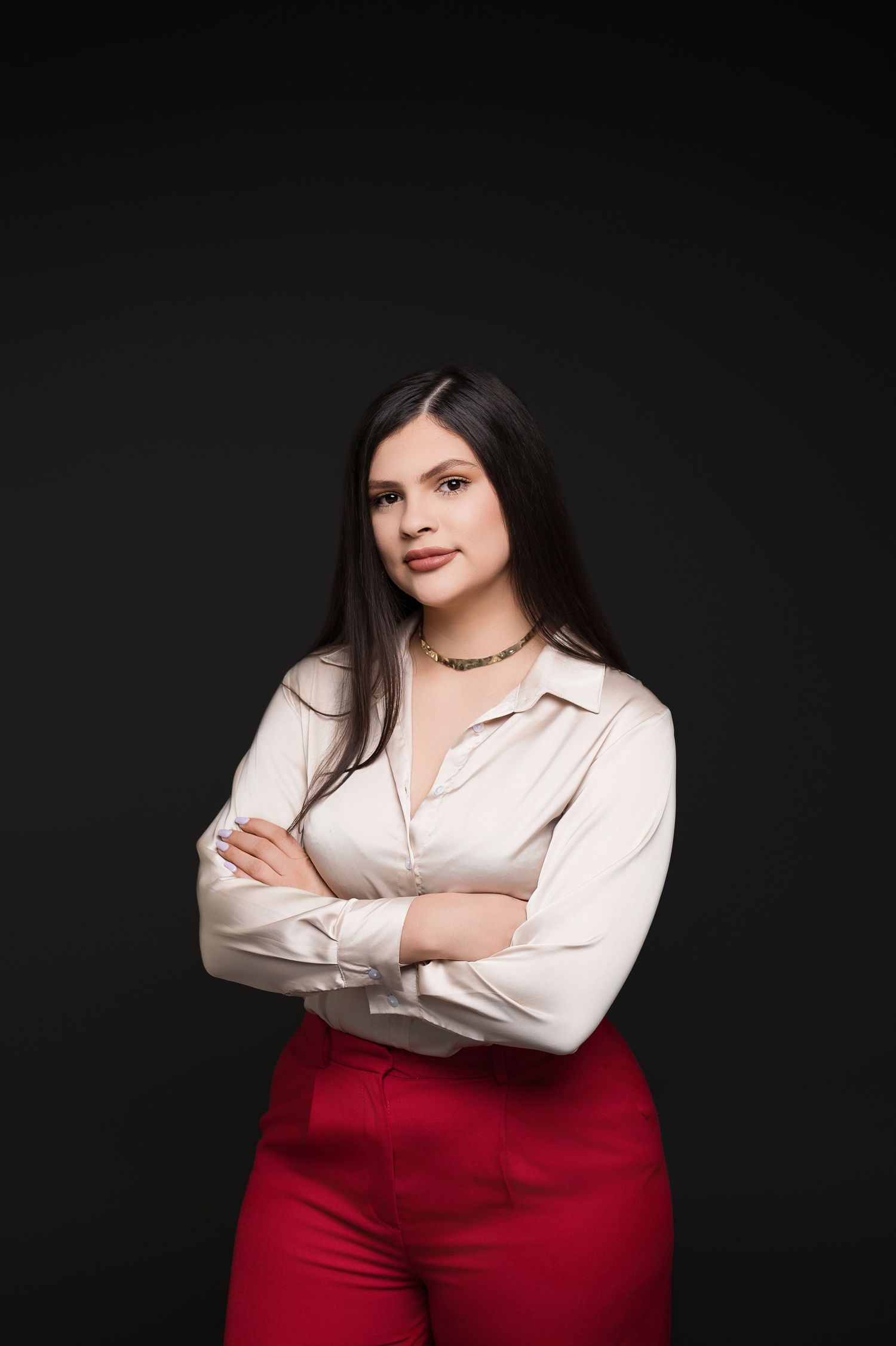 Professional studio portrait in white blouse and red pants against dark background.