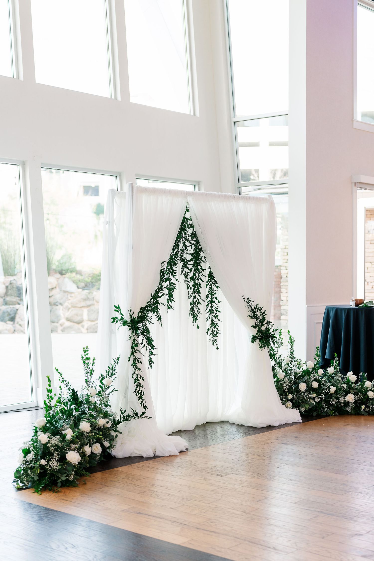 Elegant white fabric backdrop with cascading greenery and white flower arrangements for a wedding ceremony setup.