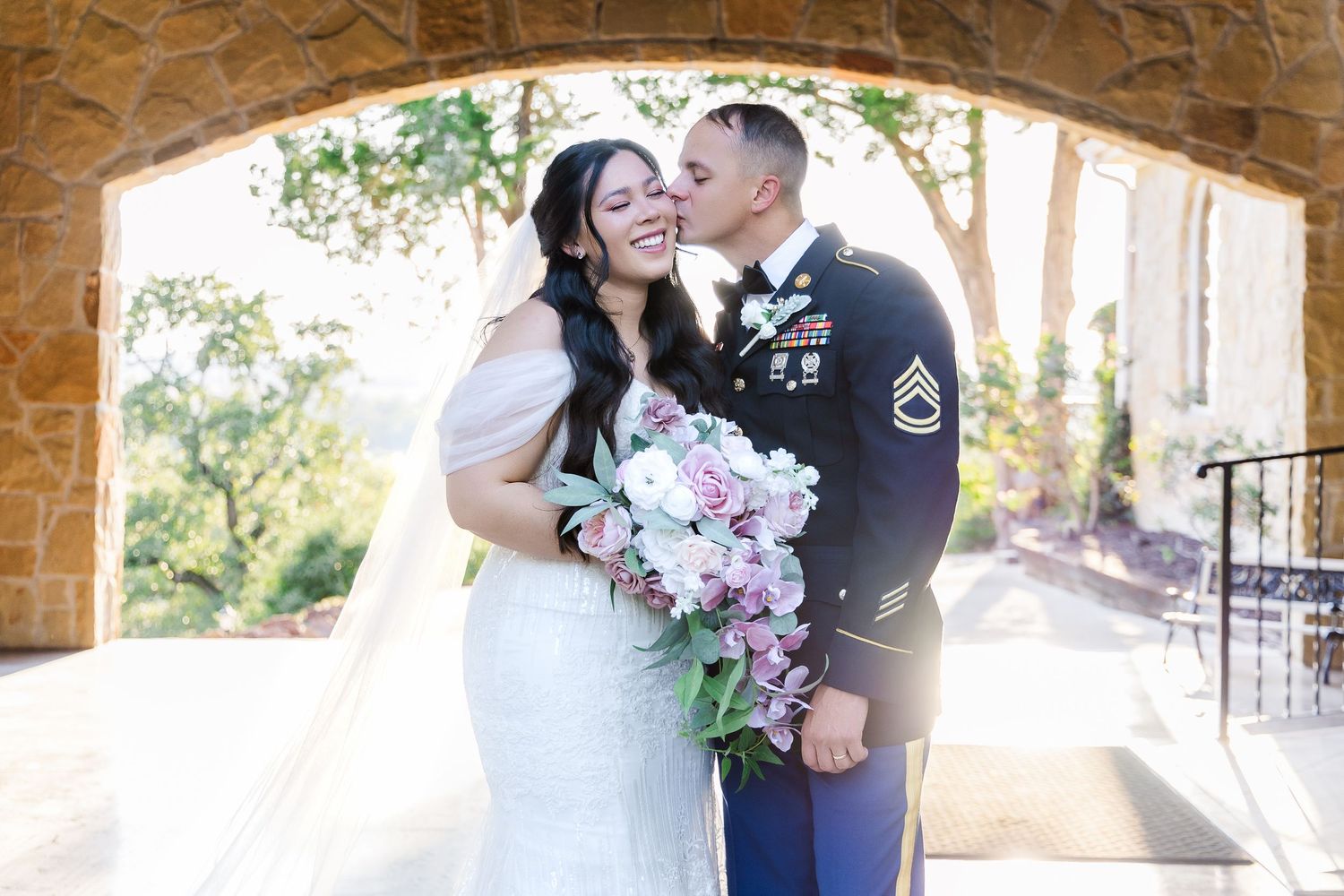 A joyful wedding moment captured under a stone archway with pink floral bouquet.