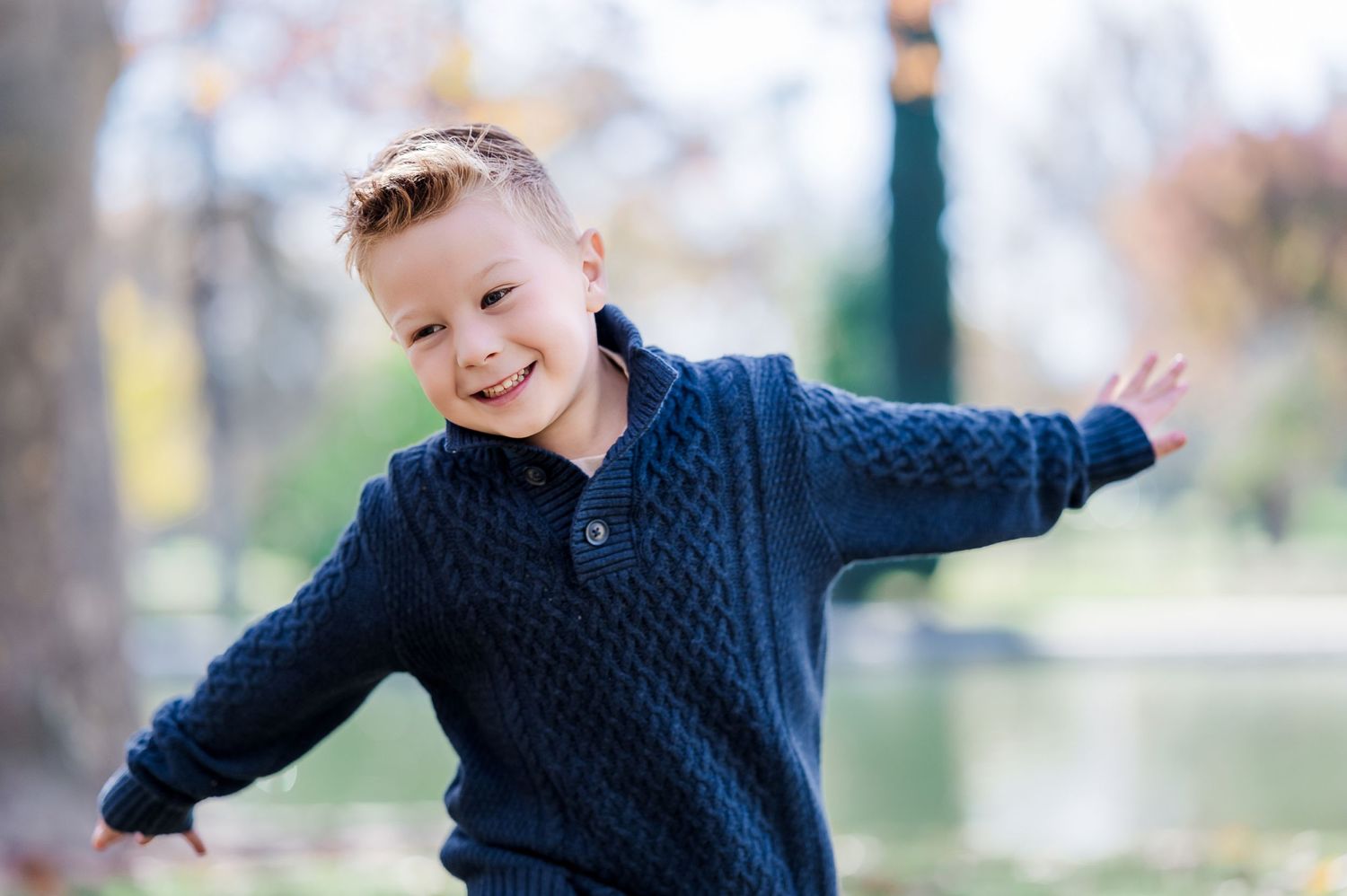 Child in navy cable knit sweater poses playfully outdoors with arms outstretched.