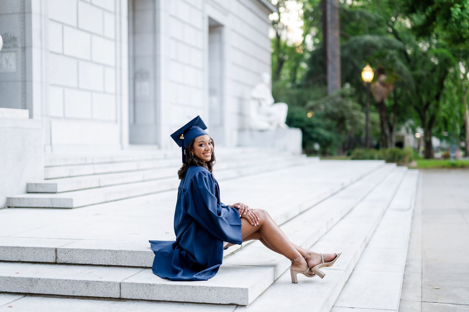 Graduate in blue cap and gown sitting on white stone steps outdoors.