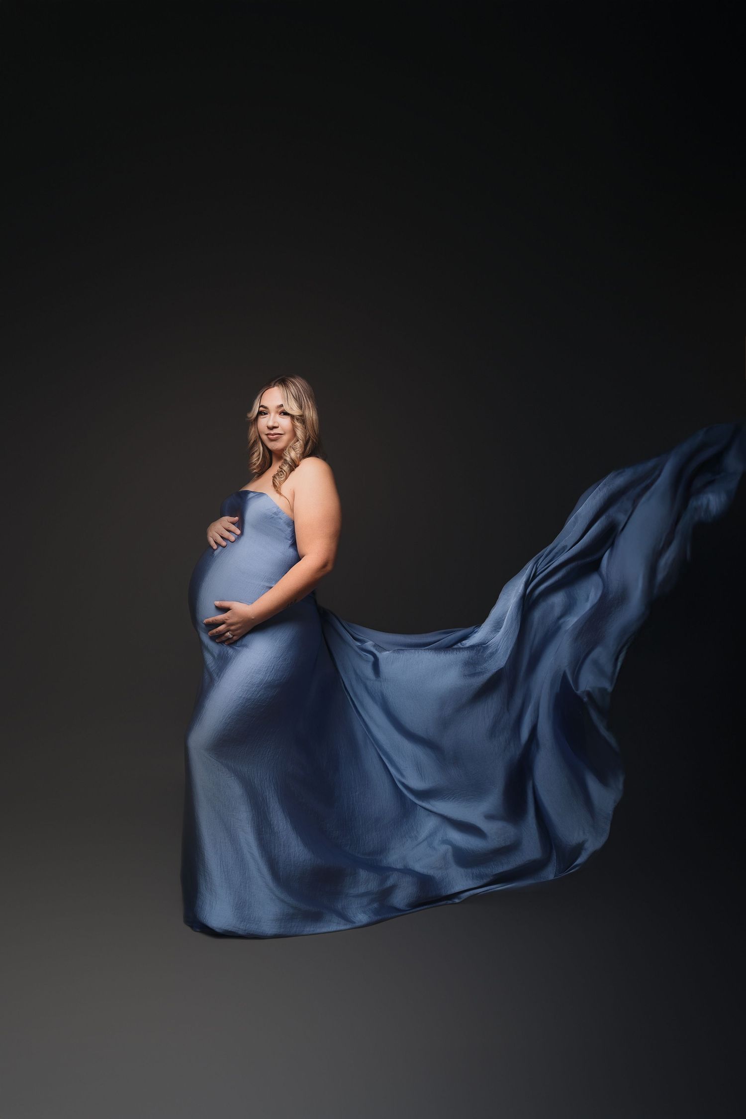 Elegant maternity session featuring blue silk fabric in motion against dark background.