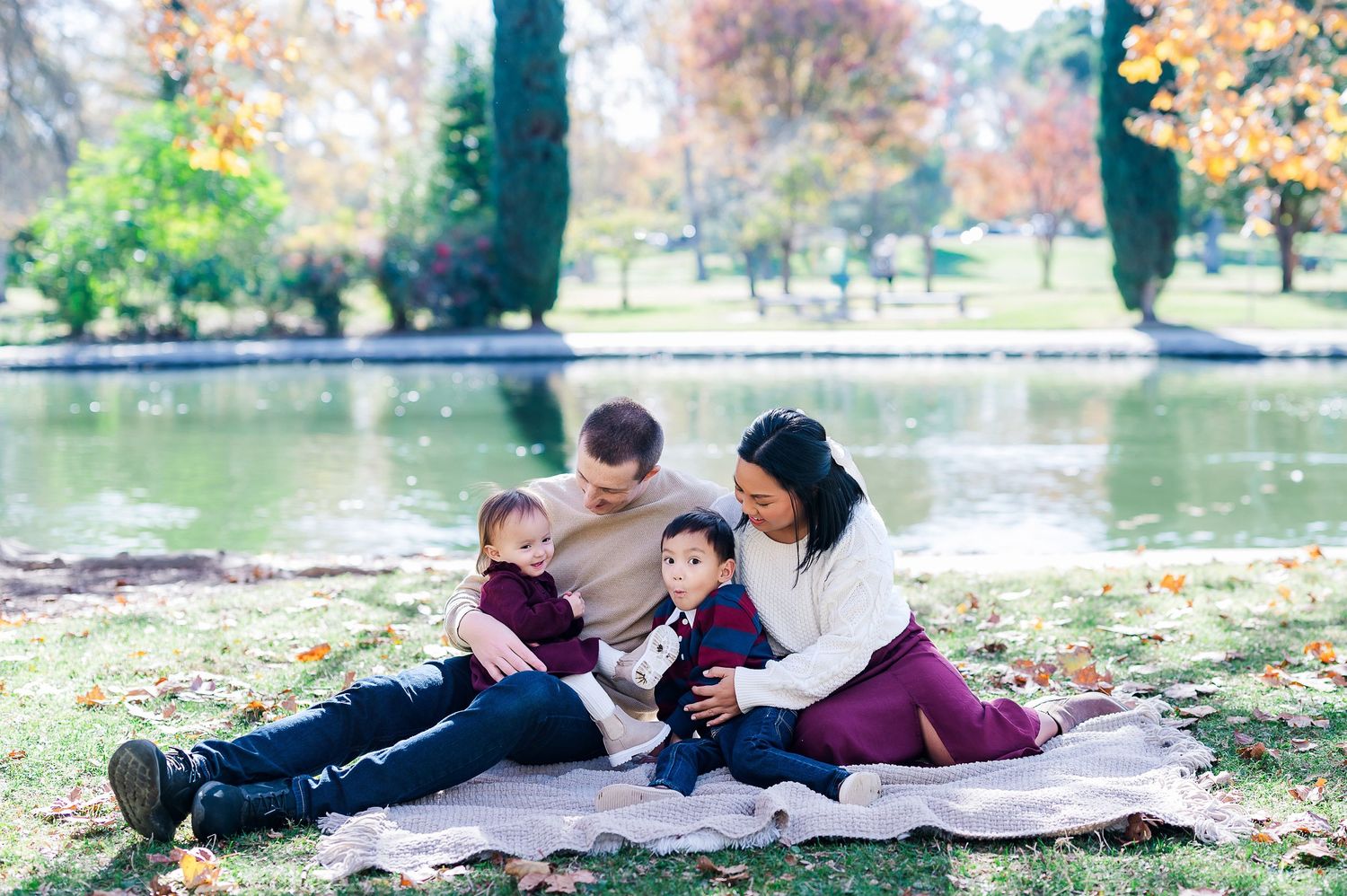 Family relaxes on picnic blanket by lake during autumn with colorful trees in background.