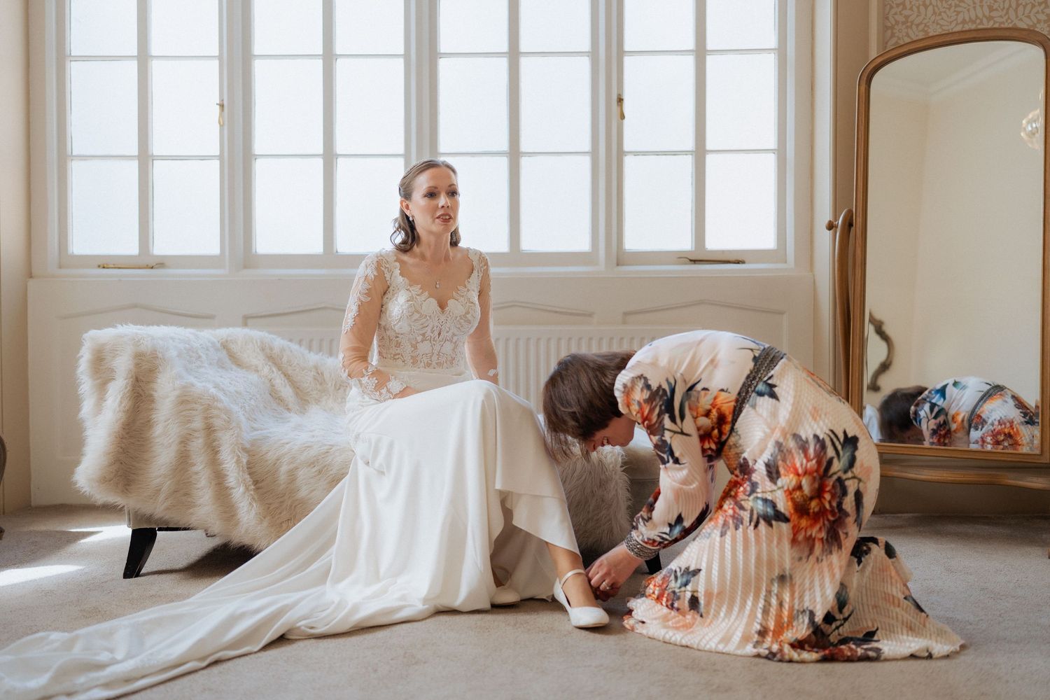 Elegant wedding dress fitting scene in sun-lit room with ornate mirror and floral arrangement. Wedding an Nonsuch Mansion