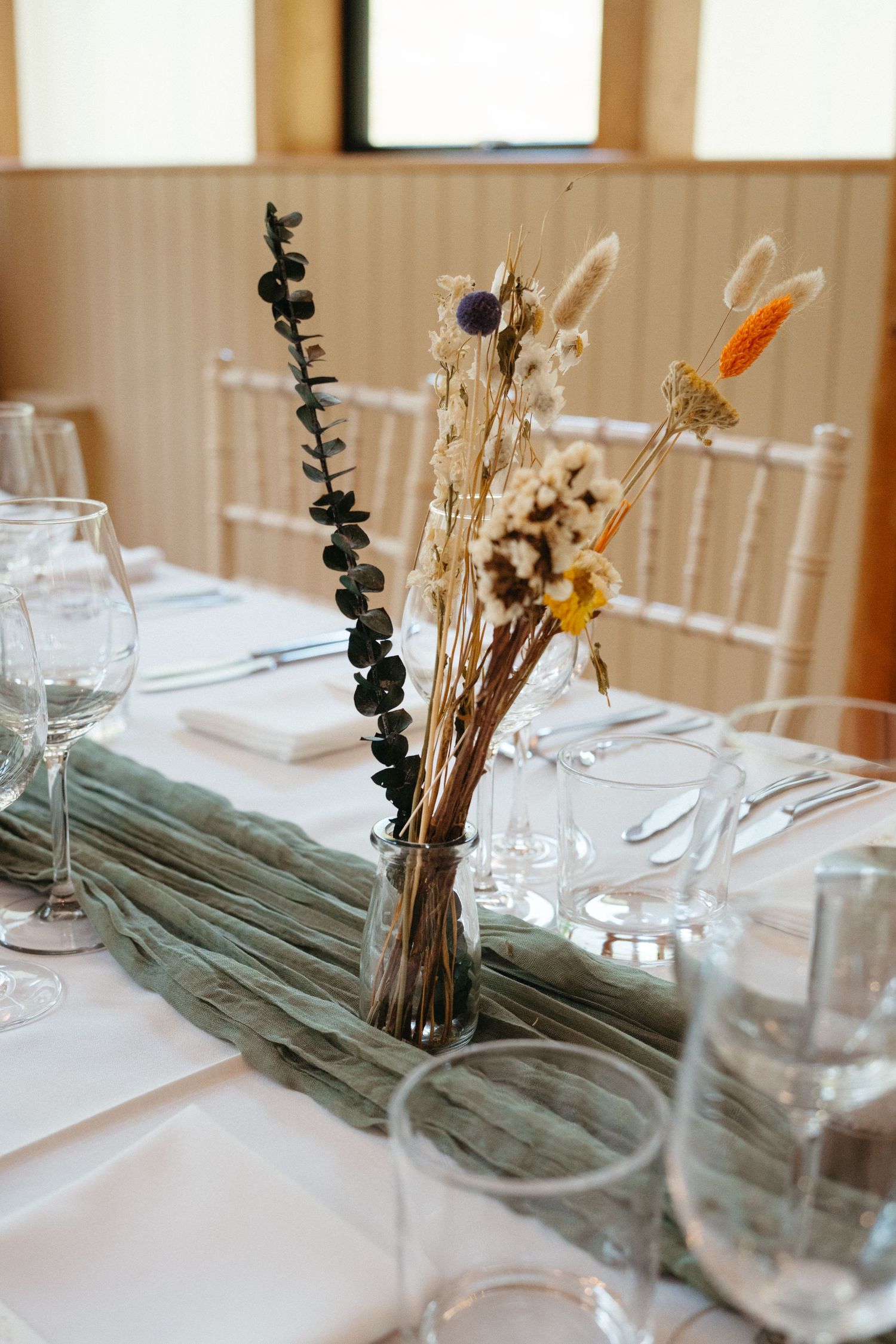 Minimalist table setting with dried floral centerpiece and wine glasses on white tablecloth at wedding reception.