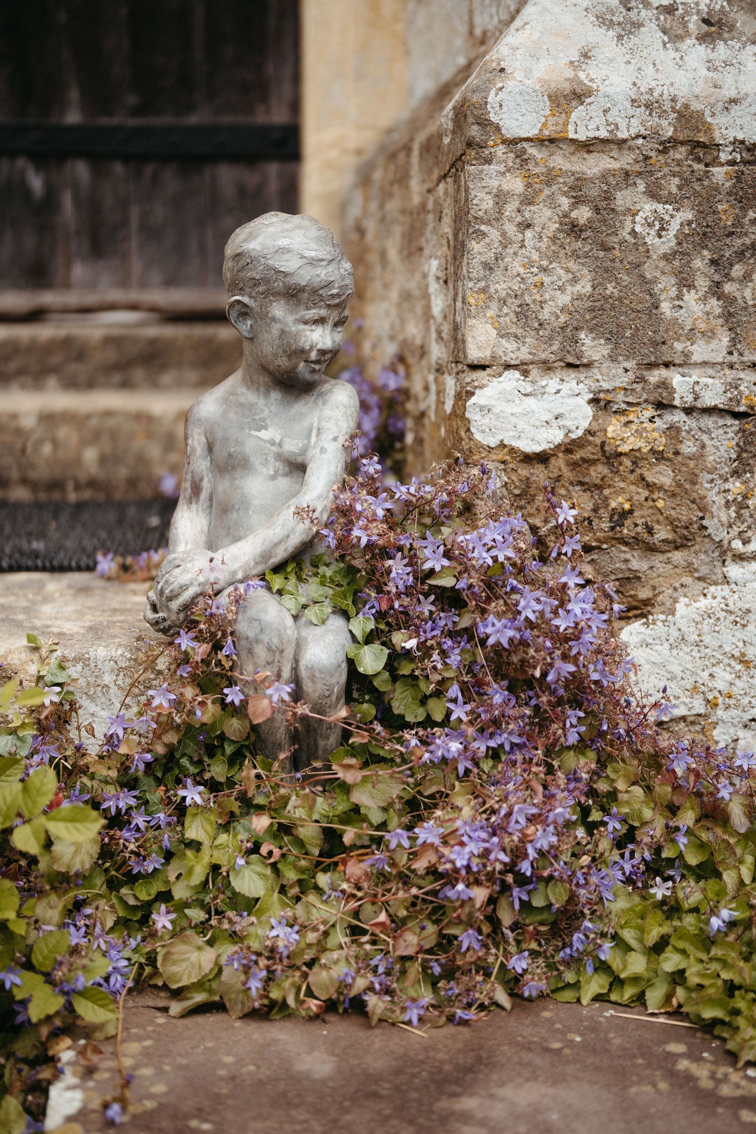 A weathered stone statue adorned with blooming purple flowers sits against an aged stone wall.