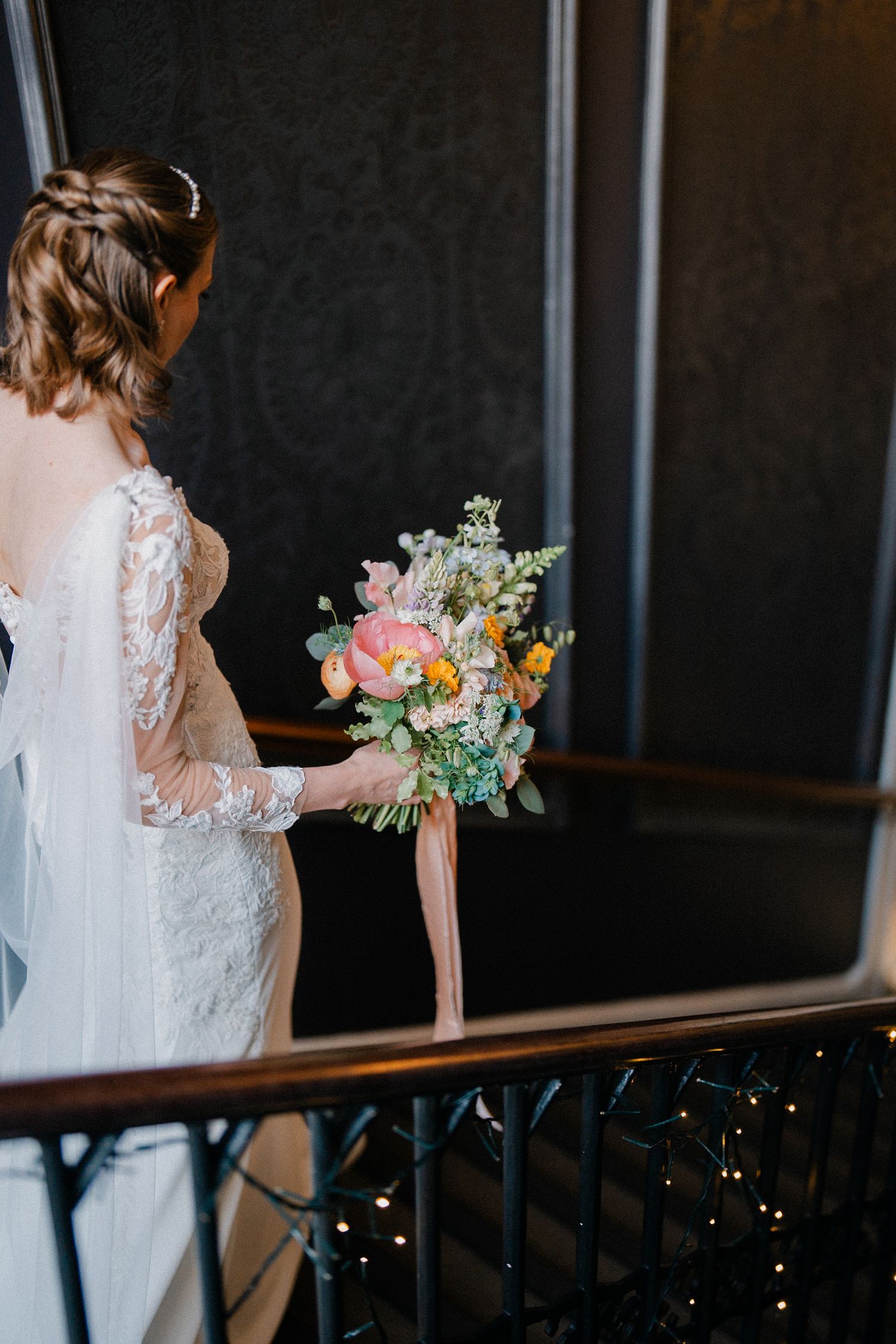 A bride in lace sleeves holding a romantic pink and white floral bouquet on a staircase. Wedding at Nonsuch Mansion