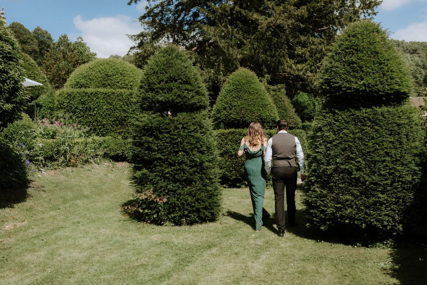 Two people stroll through a manicured garden with sculpted topiary evergreen bushes on a sunny day.