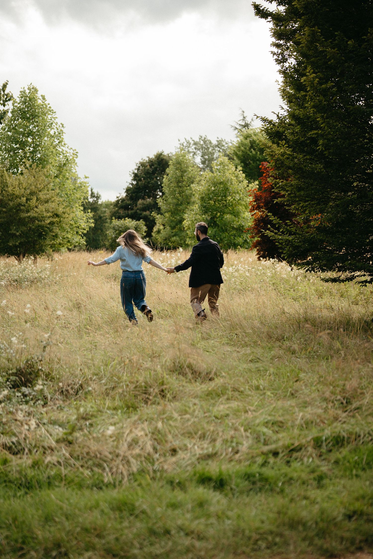 Two people run together holding hands through a meadow surrounded by autumn trees and foliage on an overcast day.