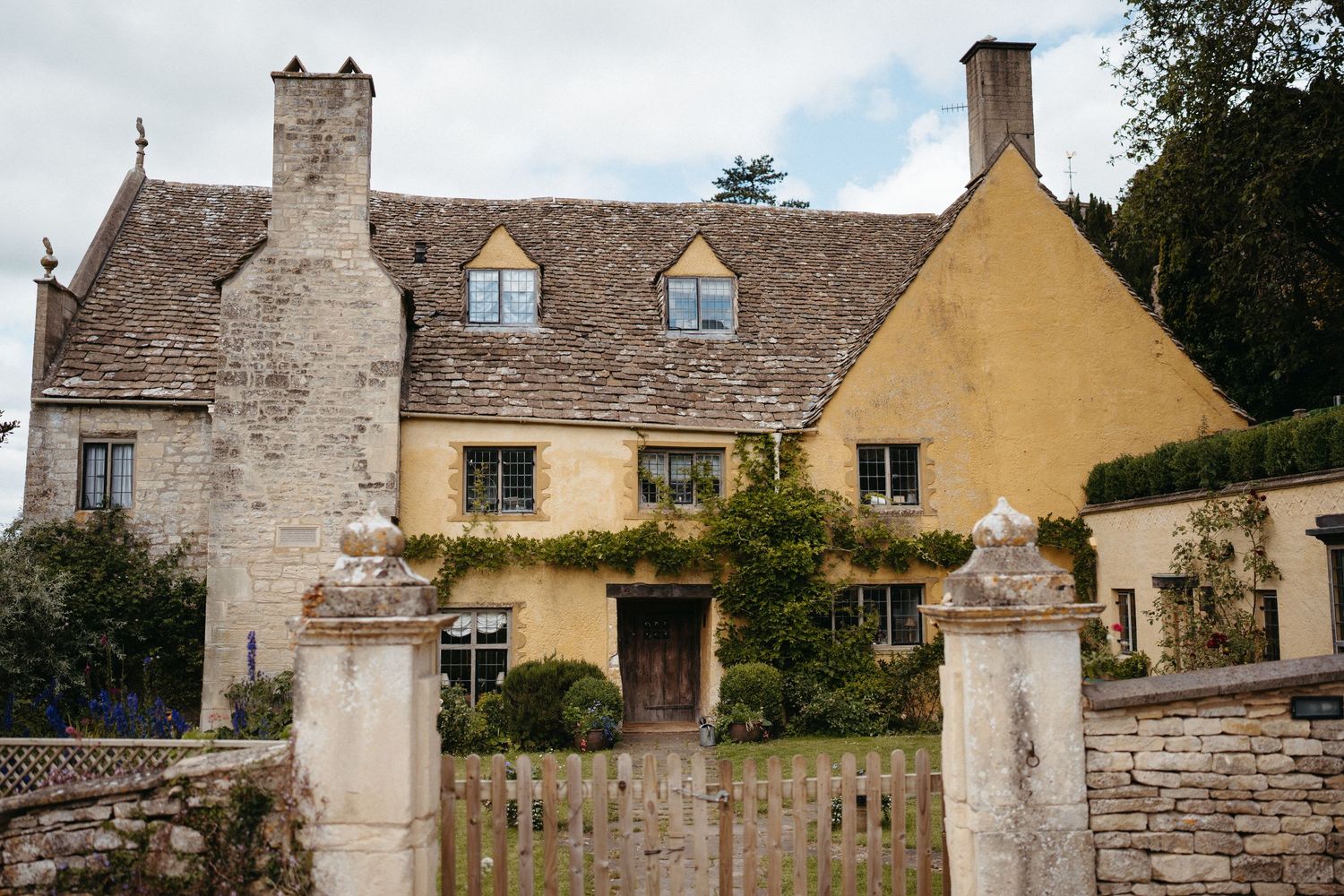 Charming Cotswold stone cottage with yellow walls and dormer windows behind stone gate posts and wooden fence.