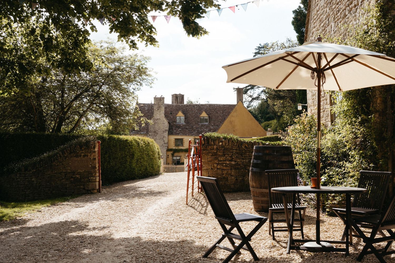 Outdoor patio seating area with umbrella and chairs overlooking garden path leading to traditional stone cottage.