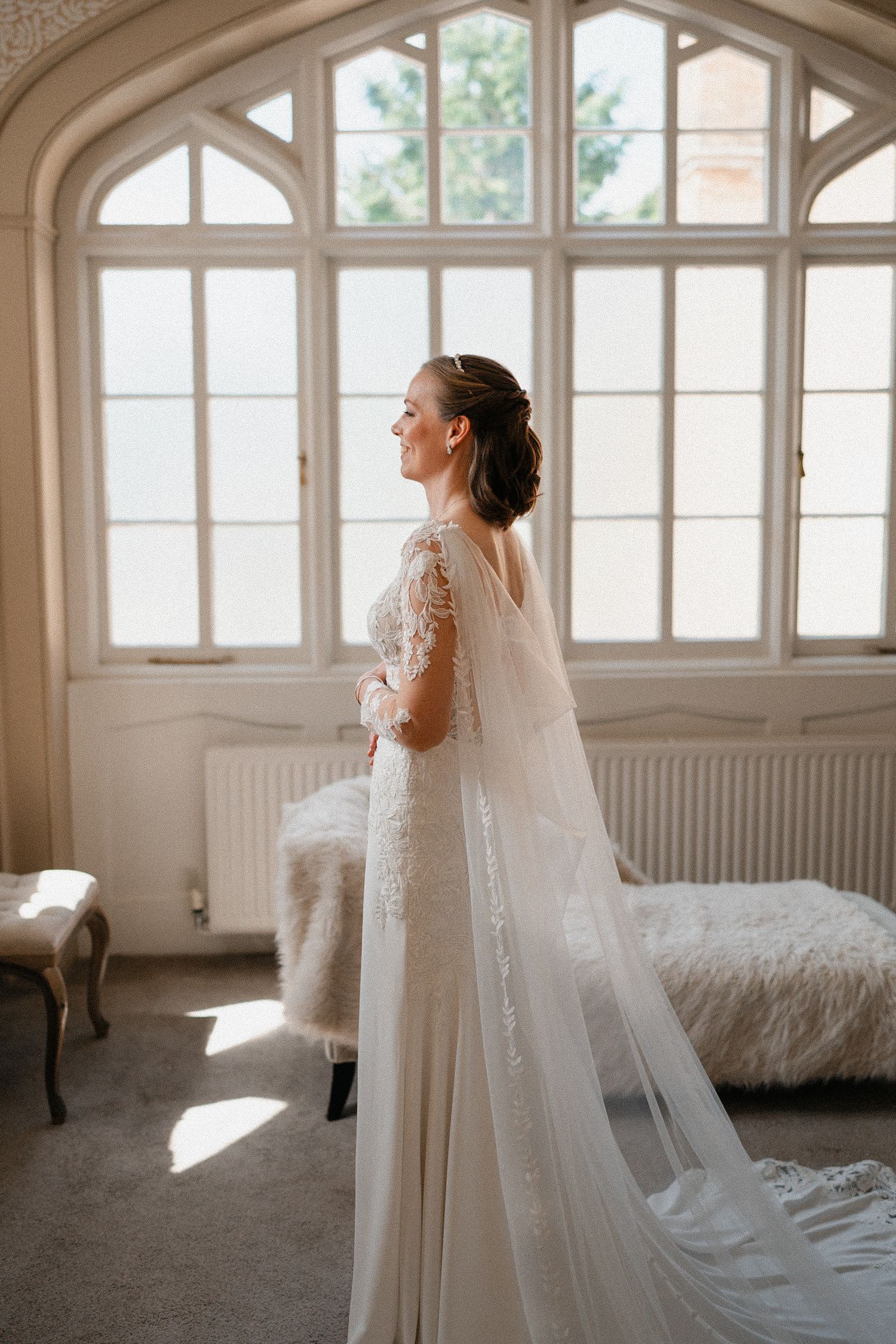 Bride in elegant white gown and veil stands in sunlit room with arched windows and vintage furniture. Wedding at Nonsuch Mansion