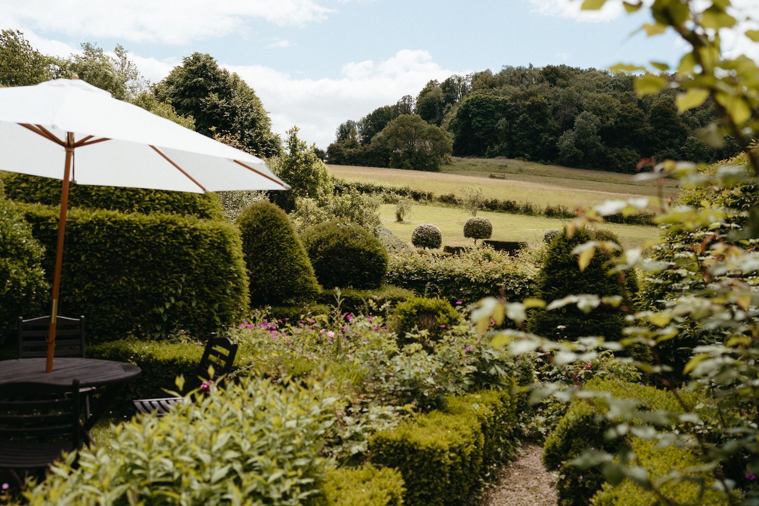 A garden patio with white umbrella overlooks rolling countryside hills and green hedges on a sunny summer day.
