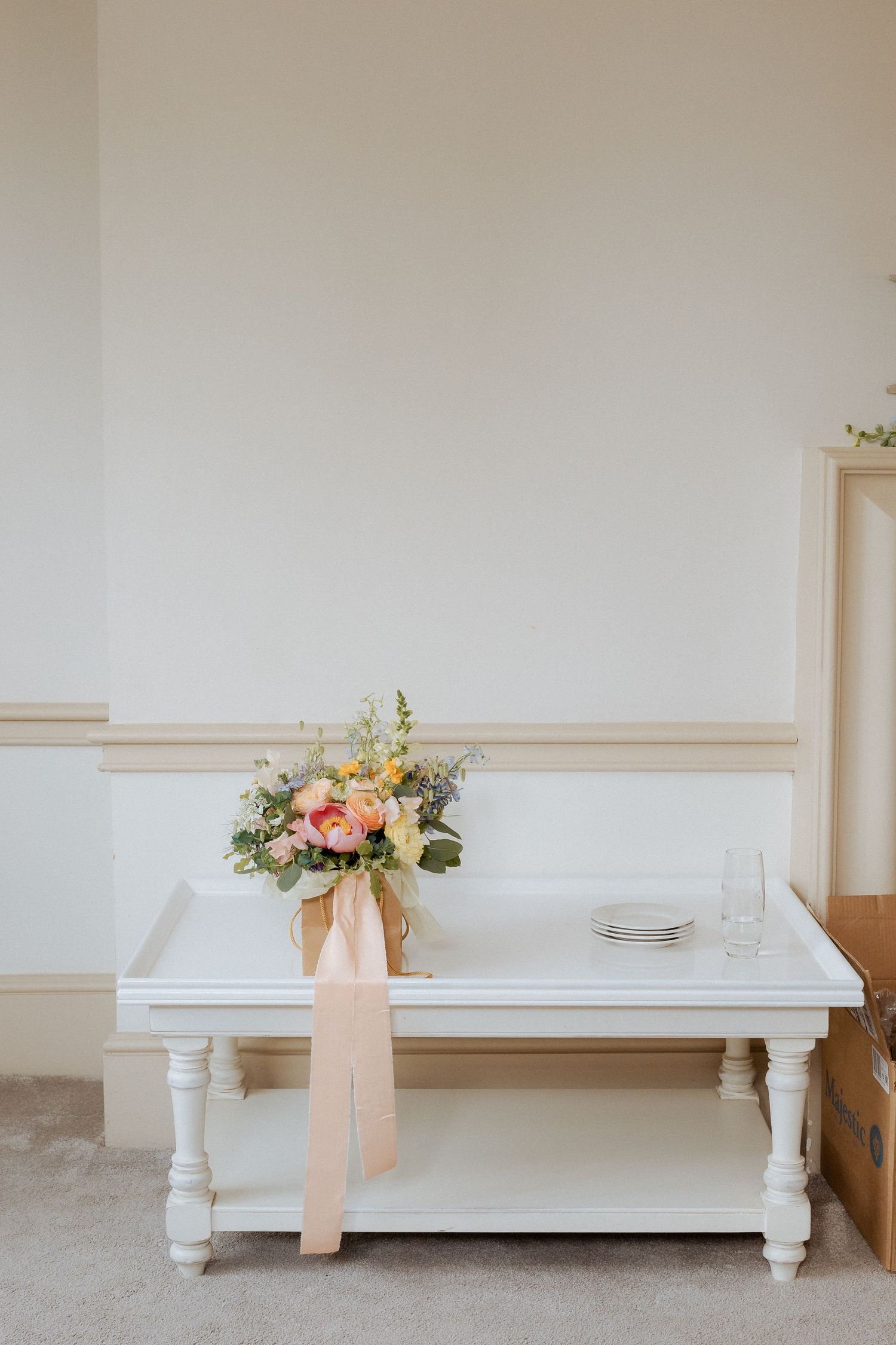 White bench against a neutral wall with delicate floral arrangements and blush ribbons creating an elegant vignette. Wedding at Nonsuch
