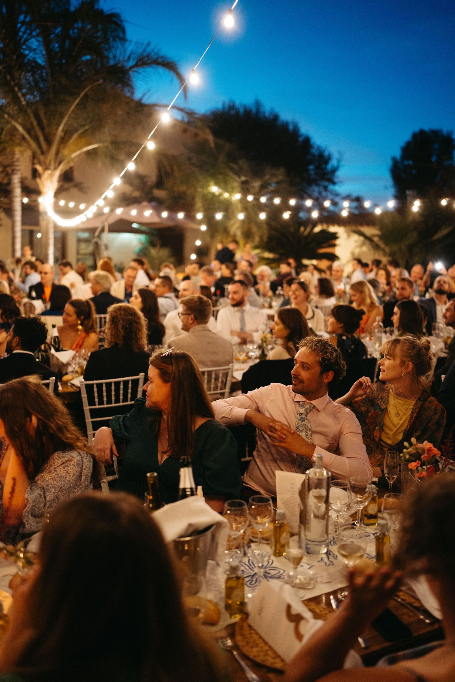 Outdoor evening wedding reception with string lights illuminating guests at dining tables.