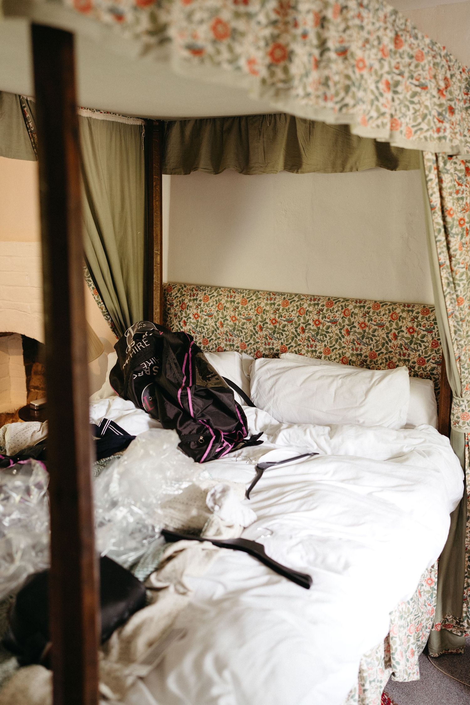 An unmade bed with white linens and a black backpack in a room with floral wallpaper and curtains.