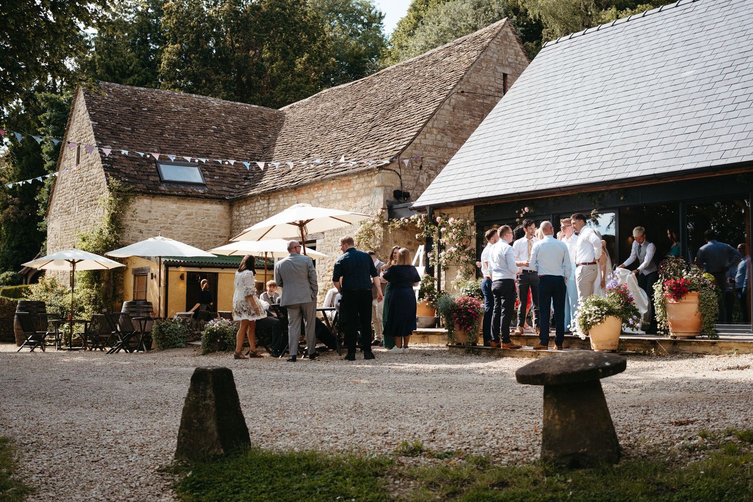 Wedding guests gather outside a rustic stone barn venue with umbrellas and outdoor seating in the courtyard.