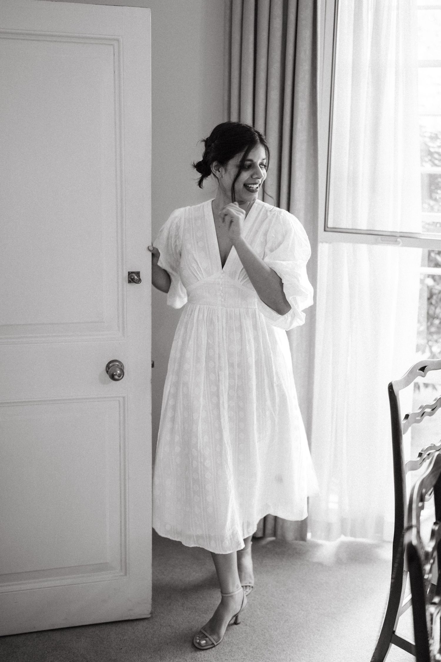 A black and white photograph of someone in a flowy white midi dress with puffy sleeves standing by a window.