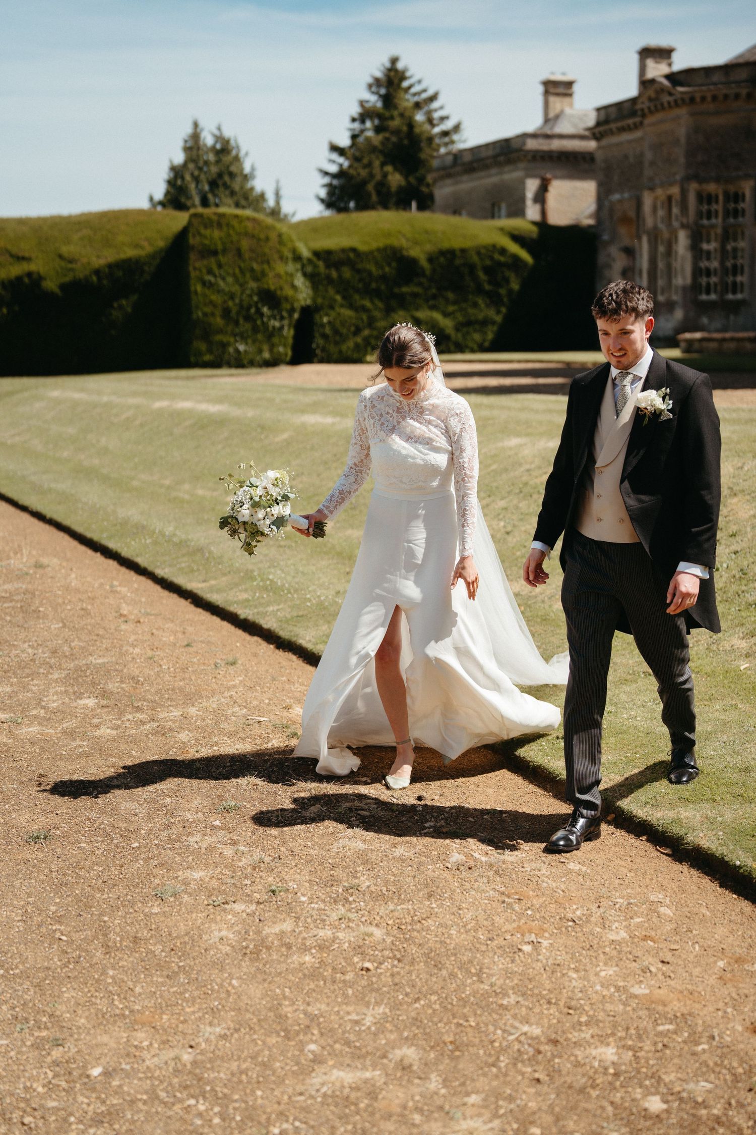 Wedding couple walking along a gravel path beside manicured hedges at an elegant estate.