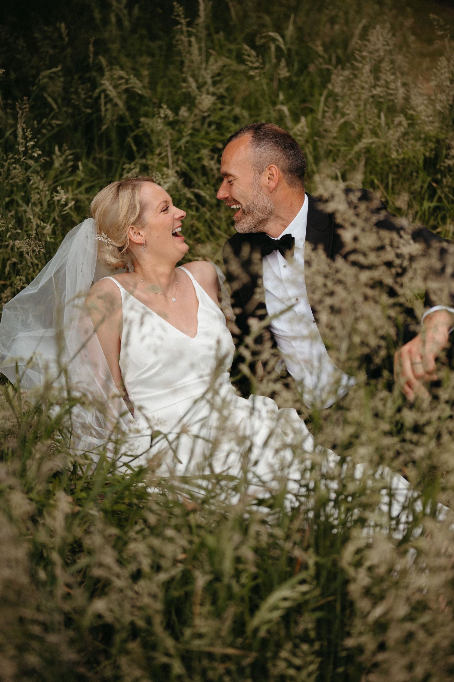 A wedding couple shares a joyful moment together in tall grass during their outdoor photoshoot.