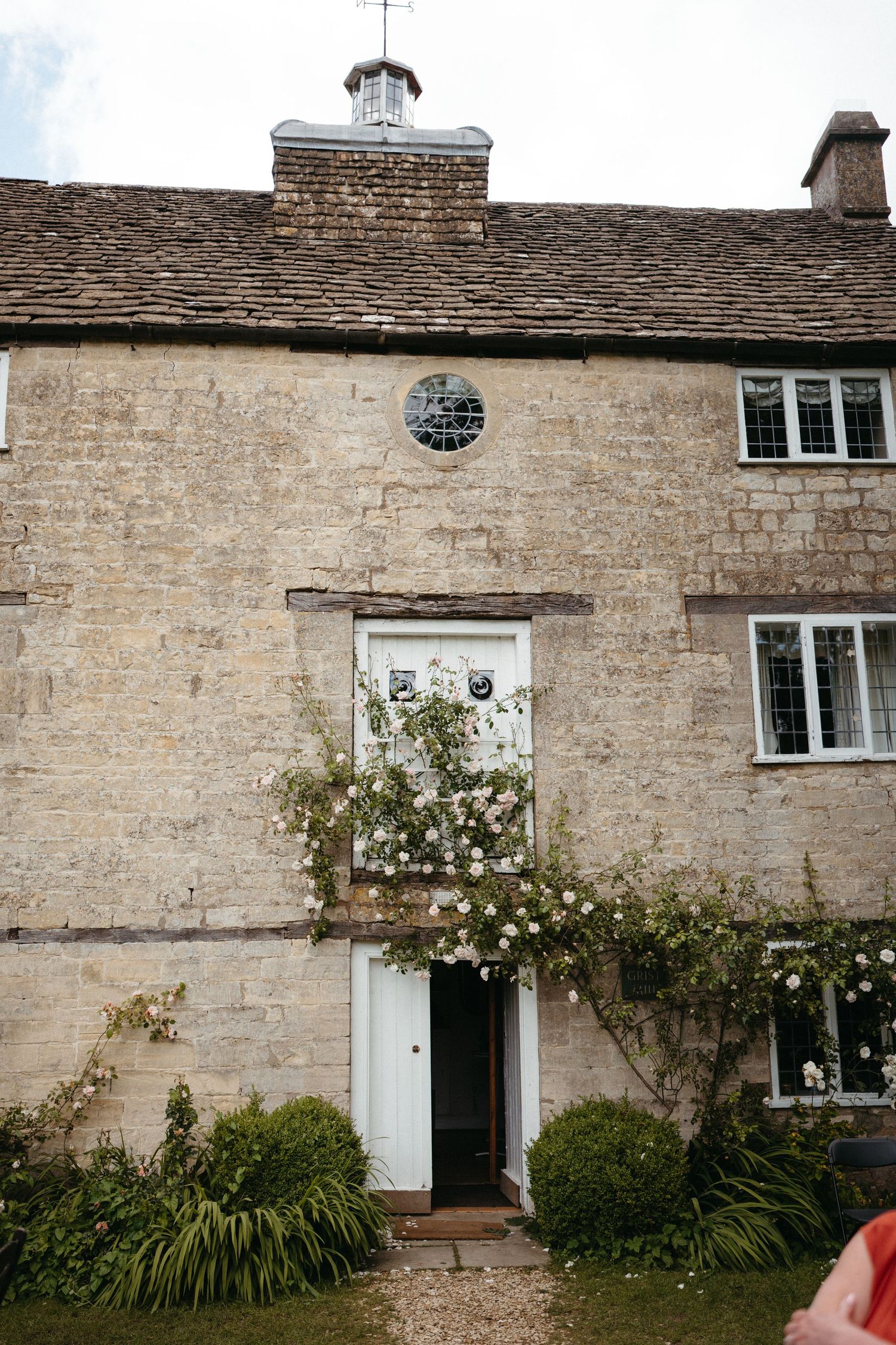 Charming stone cottage with white door, climbing roses and round plaque on exterior wall in English countryside.