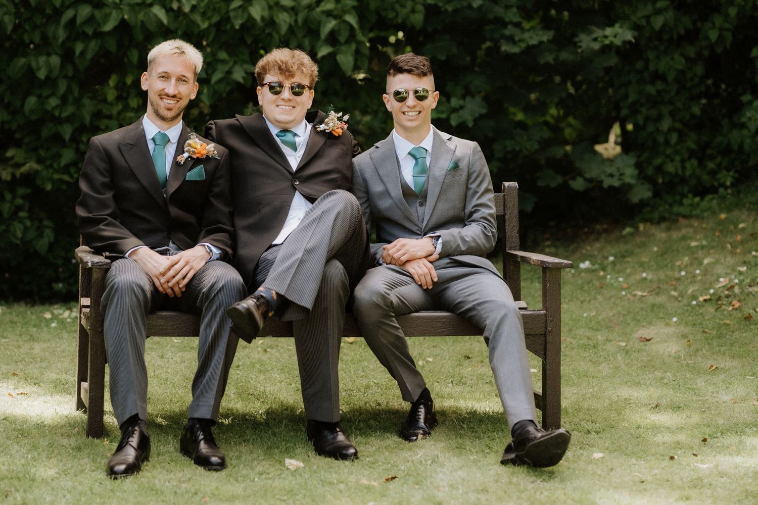 Three wedding guests in formal suits seated together on a wooden bench in a garden setting.