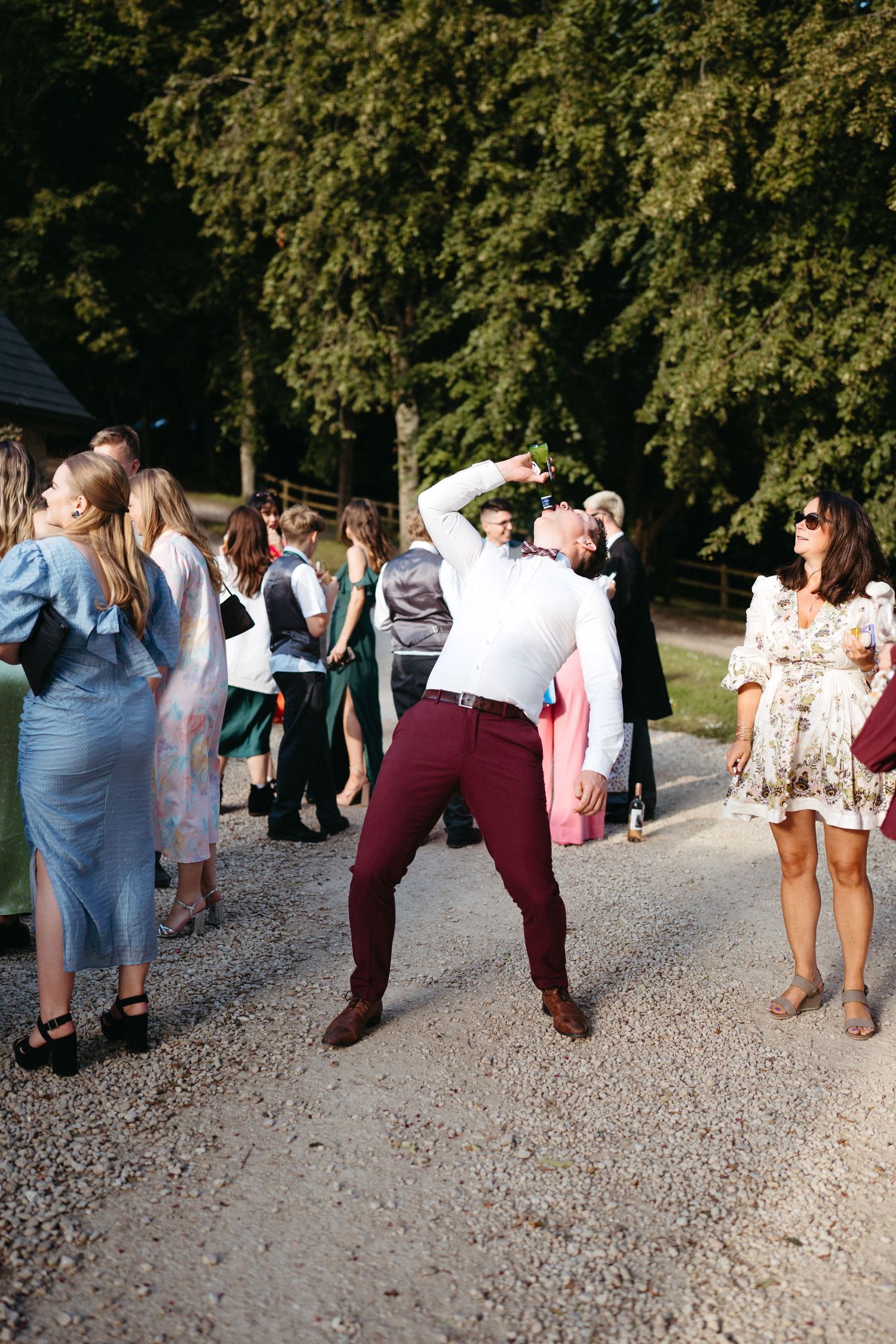 Wedding guests enjoying themselves outdoors on a gravel path surrounded by trees.