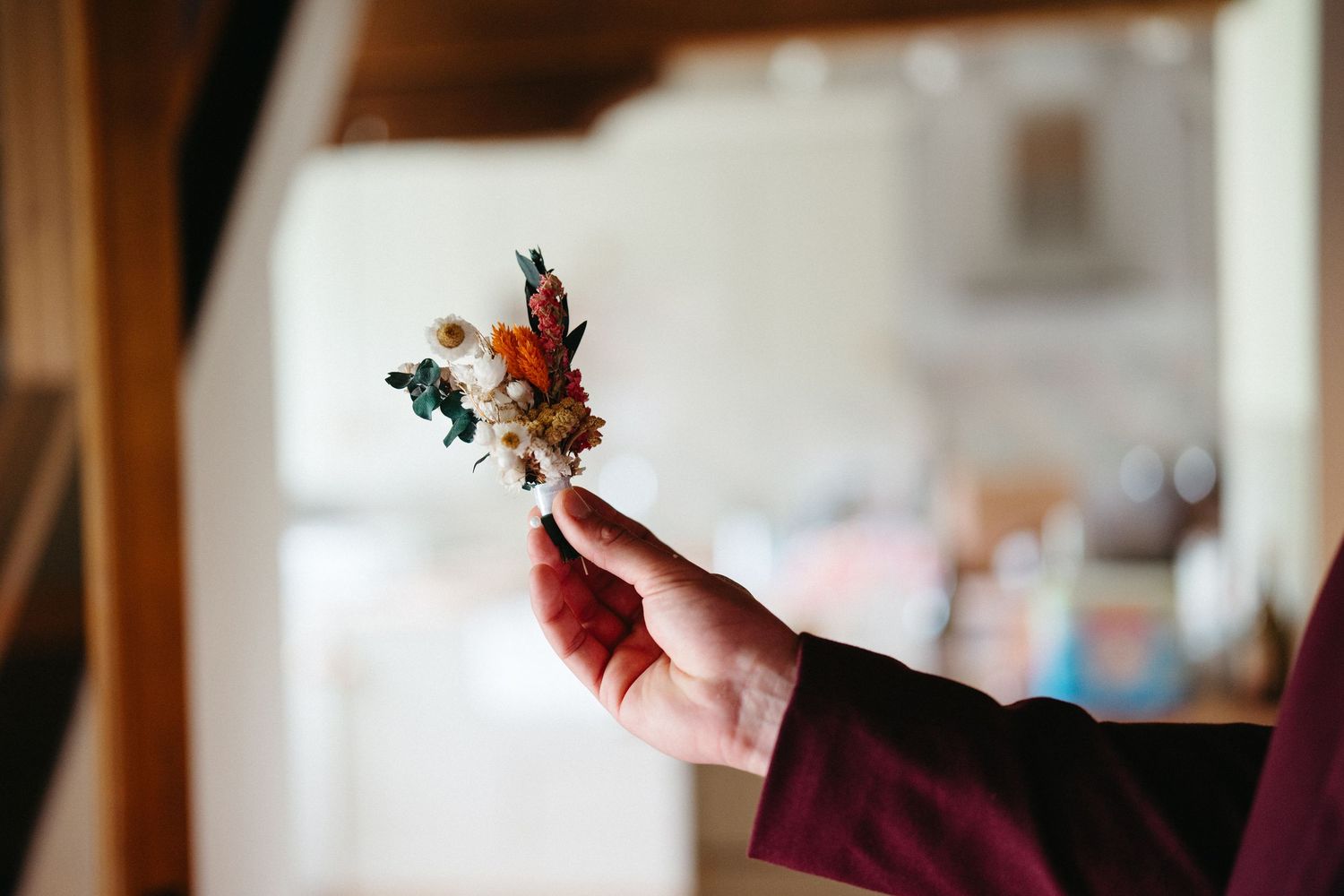 Hand in burgundy sleeve holding up a small delicate floral bouquet against a soft blurred interior background.