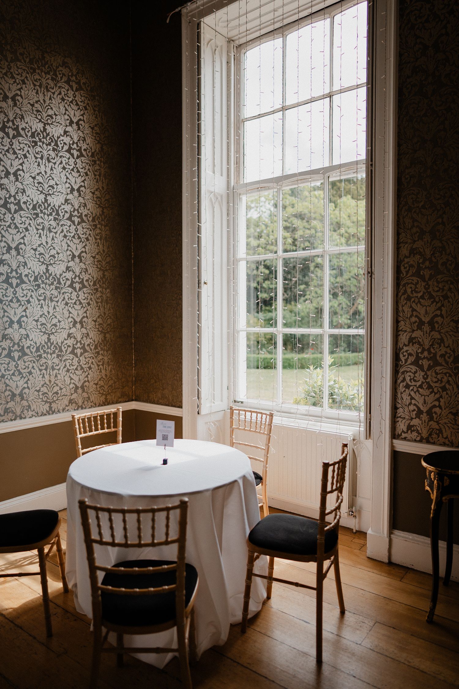 Sunny interior room with tall sash window, round table with white tablecloth, and wooden chiavari chairs.
