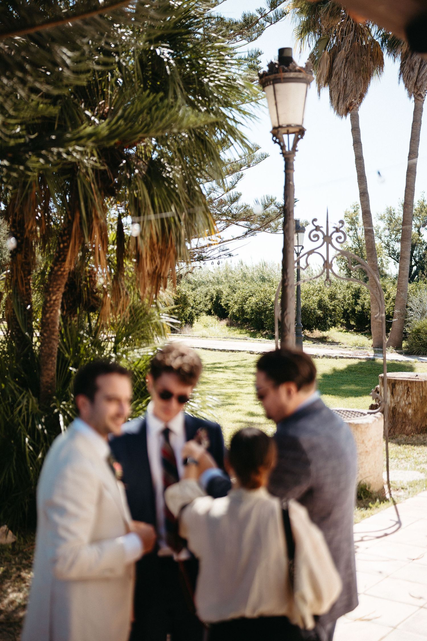 A group stands together chatting under palm trees and a vintage streetlamp in a Mediterranean garden setting.