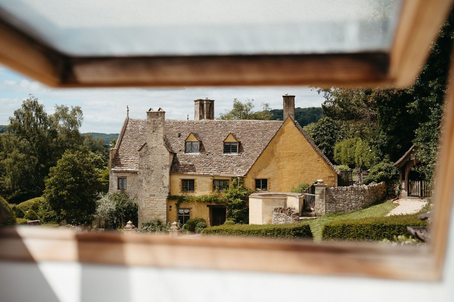 Charming traditional Cotswold stone cottage with dormer windows nestled among trees viewed through a window frame.