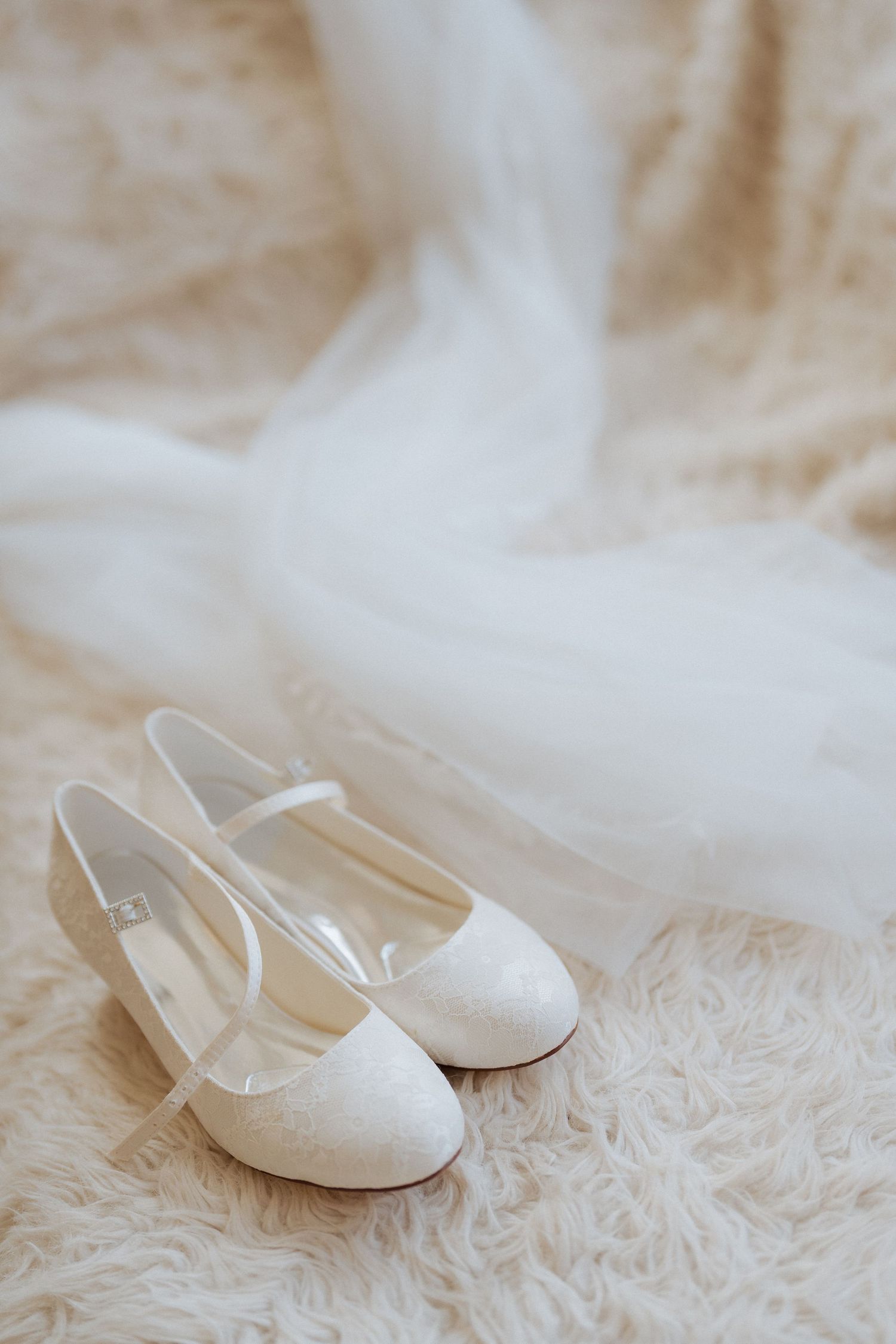 Elegant white lace bridal flats resting on a plush cream-colored fur rug with ethereal tulle fabric in the background. Wedding at Nonsuch
